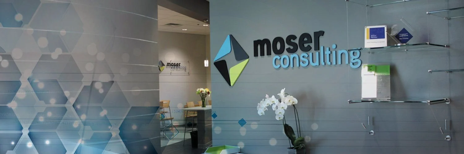 Interior of Moser Consulting office with logo on gray wall, decorative hexagon pattern, flowering plant, and reception area in background