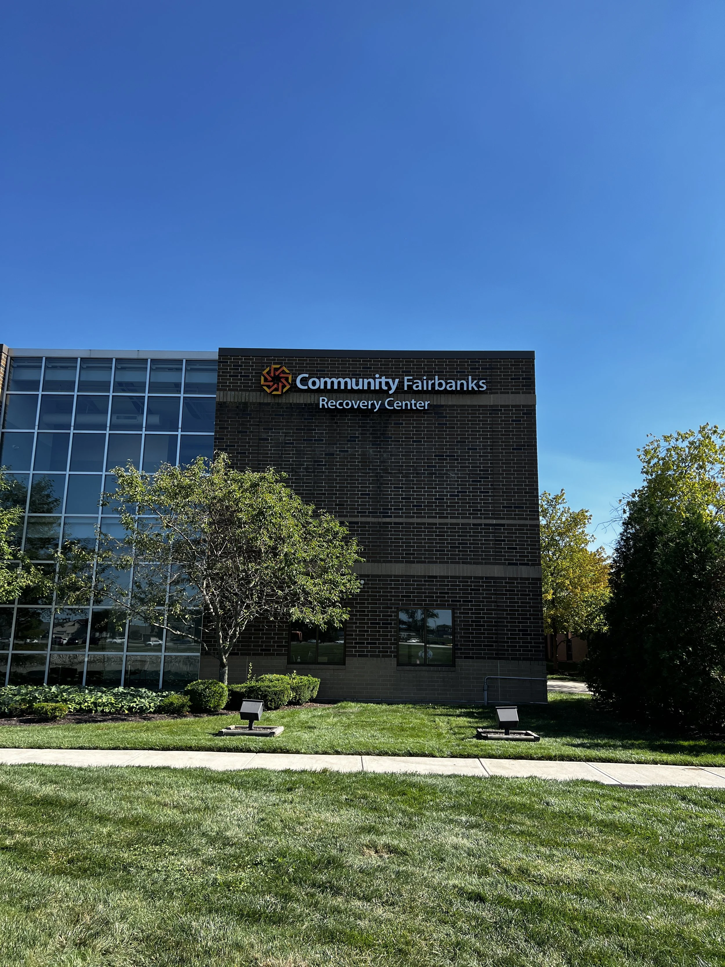 Exterior of Community Fairbanks Recovery Center building with brick and glass facade, trees, and a clear blue sky in the background.