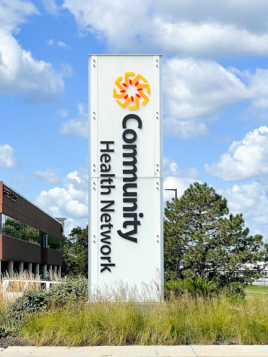 Sign reading 'Community Health Network' with a logo, on a white background, under a partly cloudy blue sky, with trees and grass nearby.