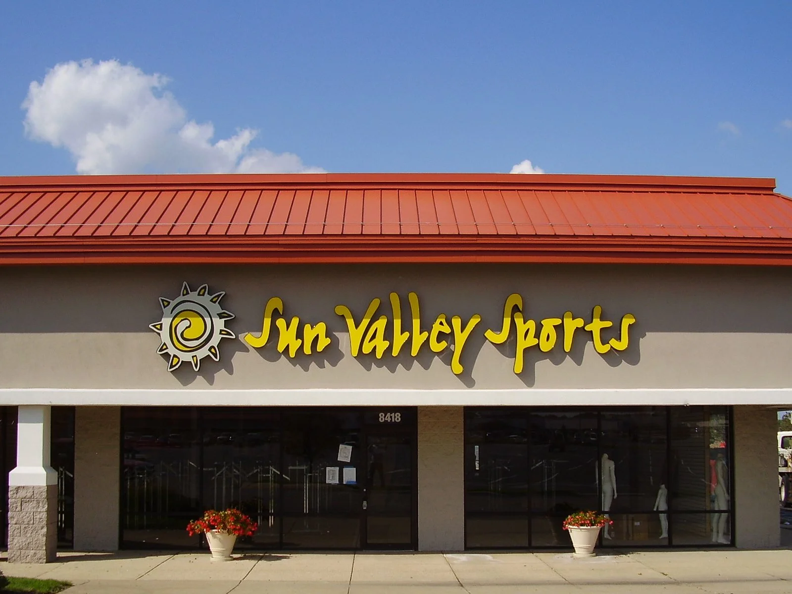 Sun Valley Sports storefront with a yellow logo and text on a beige building, a red roof, and potted plants outside, under a blue sky with clouds.