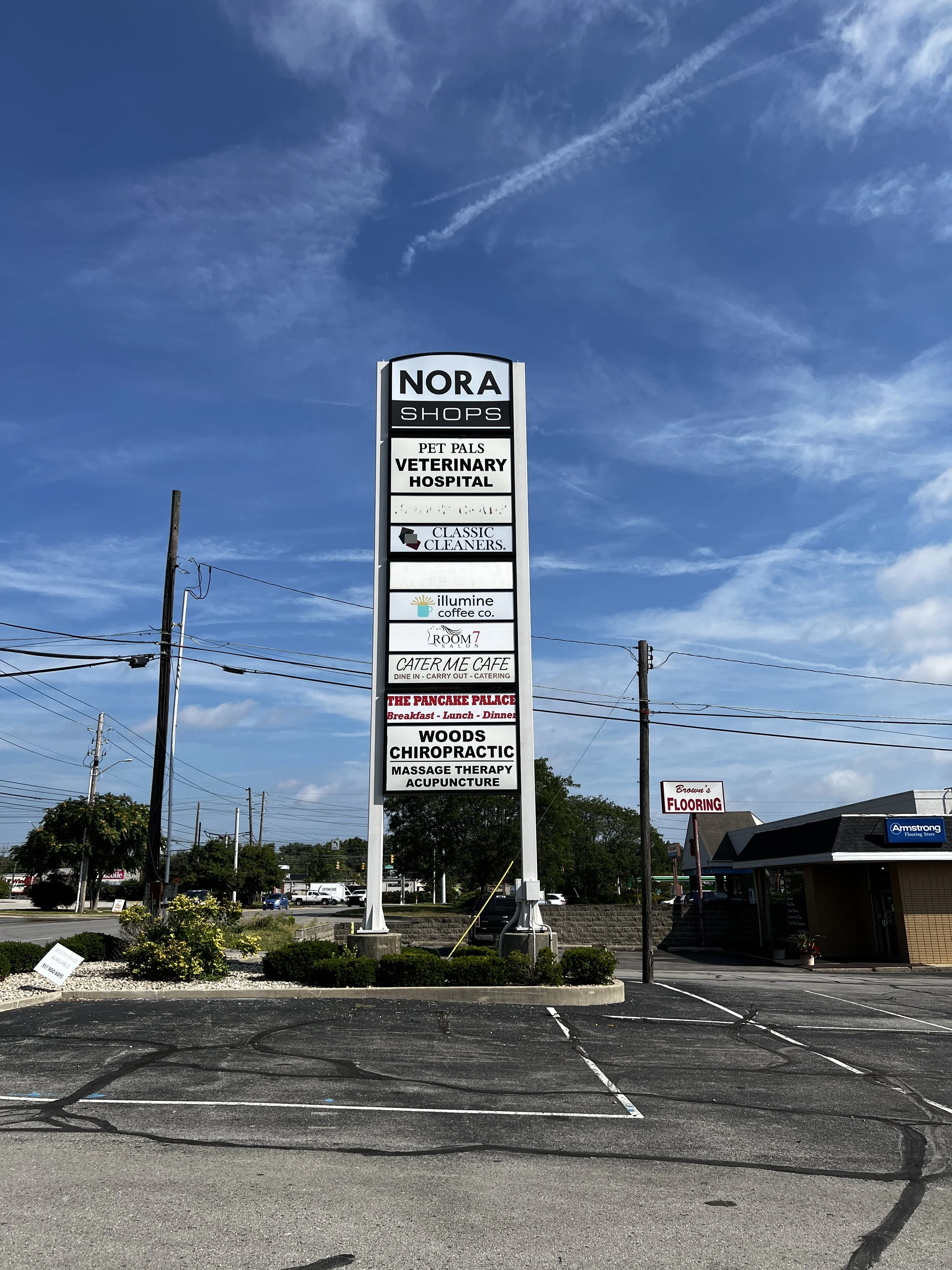 A tall outdoor sign listing various businesses including Nora Shops, Pet Pals Veterinary Hospital, Classic Cleaners, Illumine Coffee Co., Room 7 Salon, Cater Me Cafe, The Pancake Palace, Woods Chiropractic, Massage Therapy, and Acupuncture. The sign 