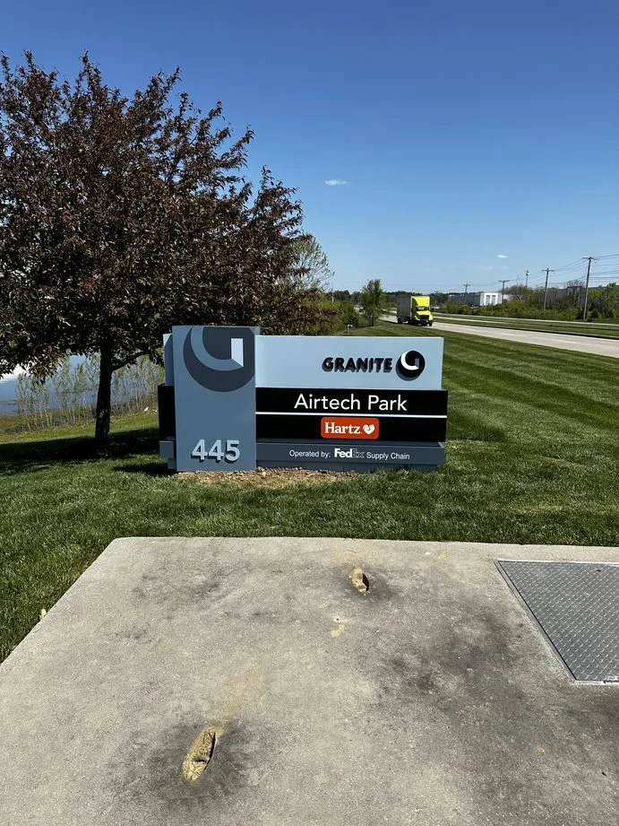 Sign for Granite Airtech Park with lot number 445, operated by FedEx Supply Chain, and sponsored by Hartz. There is a tree, a grassy area, road, and a truck in the background.