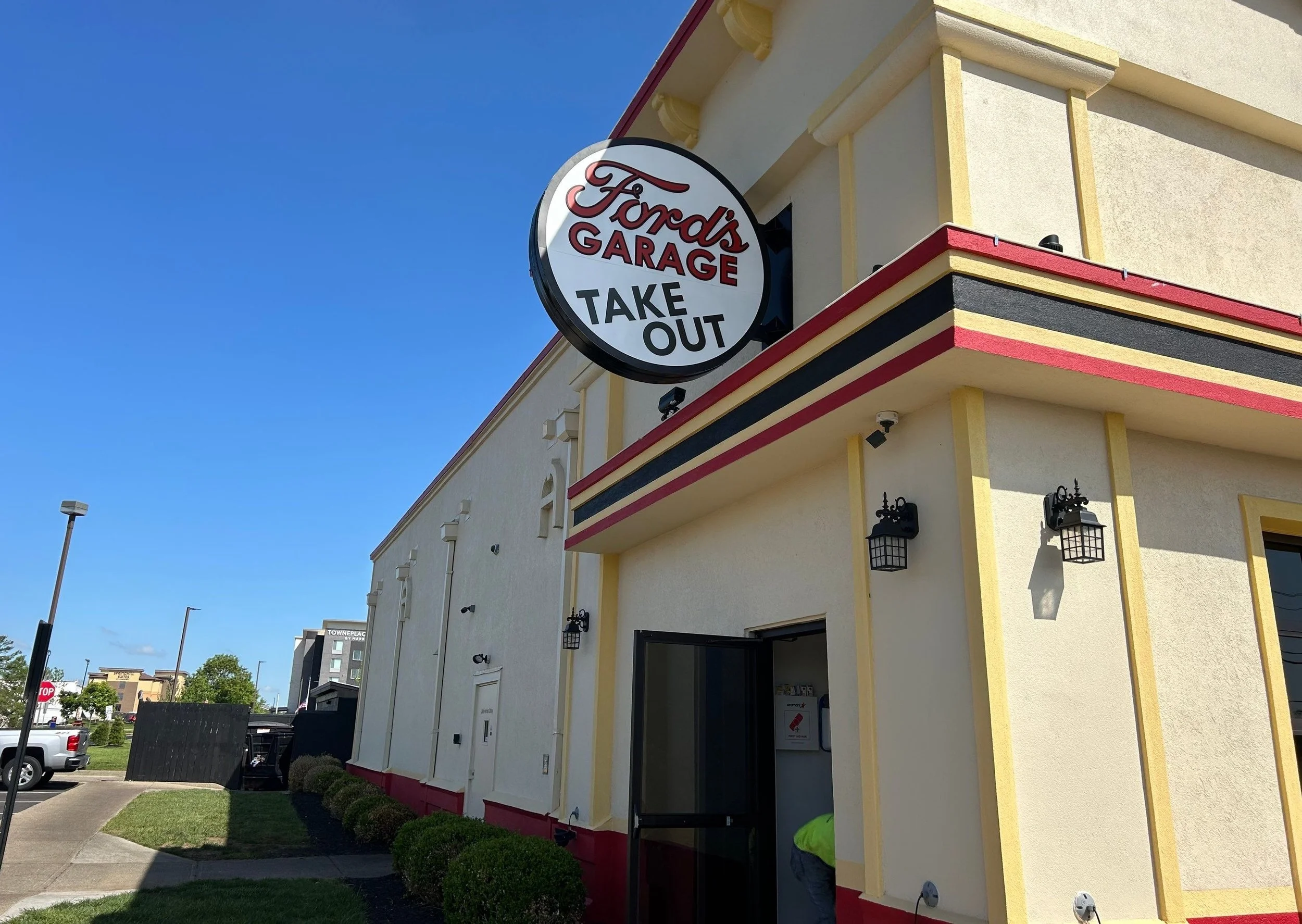 Exterior of a building with a large round sign that reads "Ford's Garage Take Out" in red and black letters. The building is painted light yellow with red and black trim, and there are outdoor lantern-style light fixtures on the wall. The sky is clea