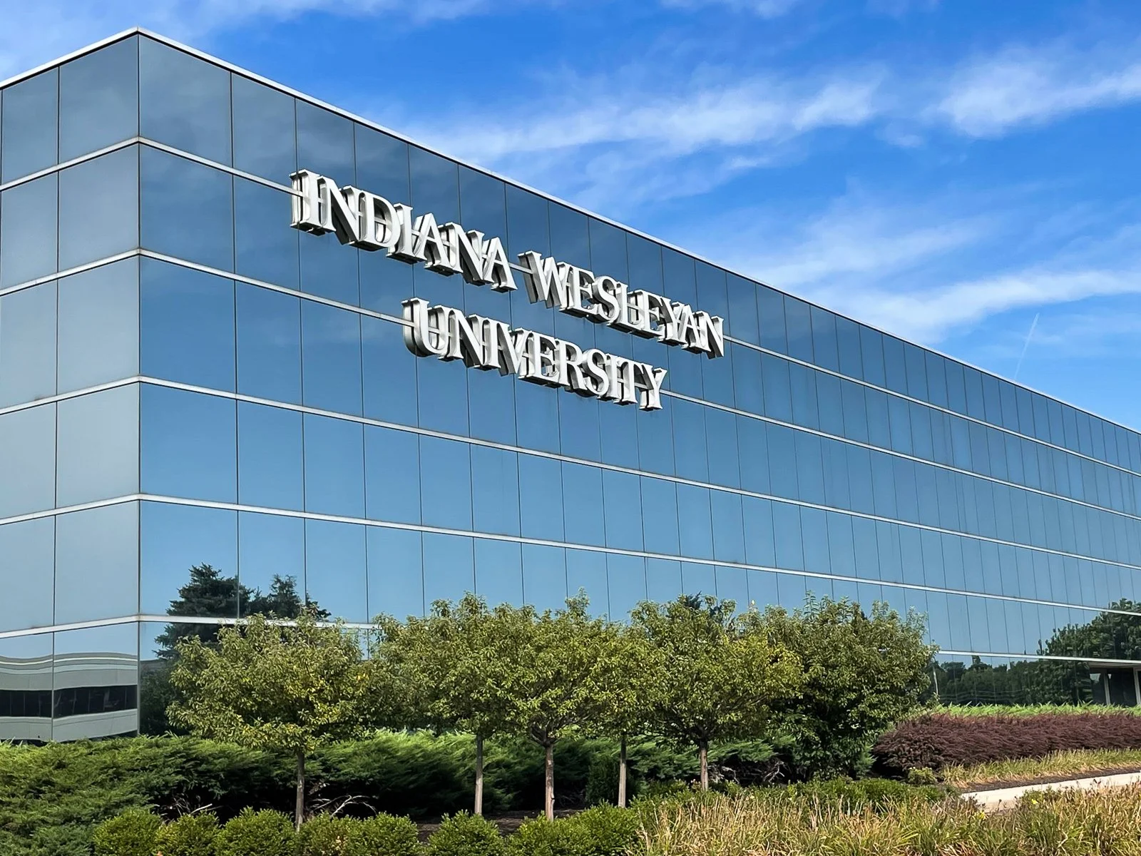 Exterior view of Indiana Wesleyan University building with reflective glass windows, and the university's name on the side, surrounded by trees and landscaping, under a partly cloudy blue sky.