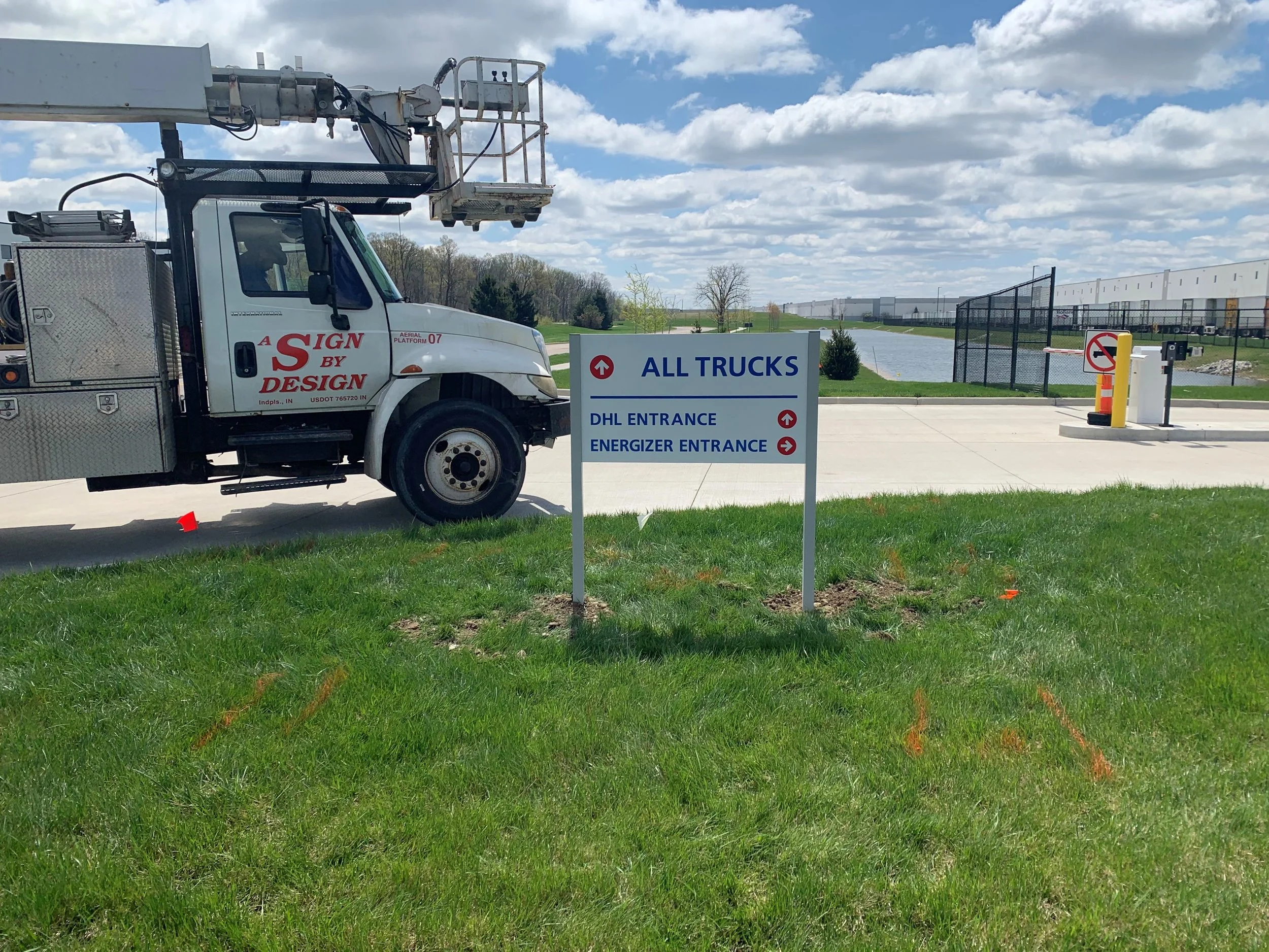 A white truck with a hydraulic lift parked near a sign that says 'All Trucks' with arrows pointing to entrances labeled 'DHL Entrance' and 'Energizer Entrance.' There is a grassy area and a body of water with a fence and industrial buildings in the b