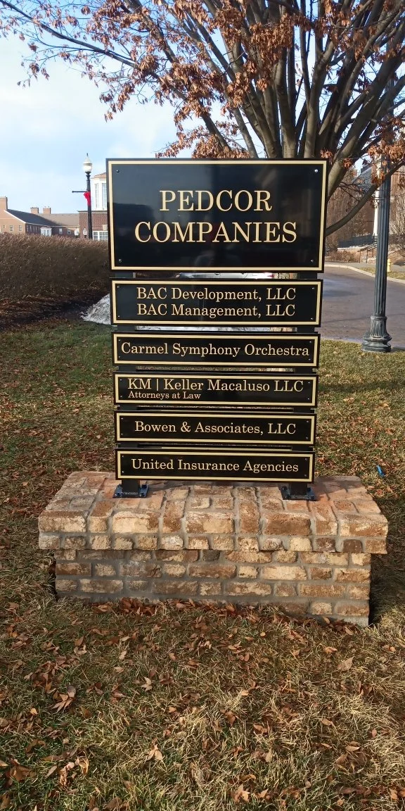 Sign for Pedcor Companies featuring names of various business entities including BAC Development, LLC, BAC Management, LLC, Carmel Symphony Orchestra, KM | Keller Macaluso, LLC, Bowen & Associates, LLC, and United Insurance Agencies, situated on a br