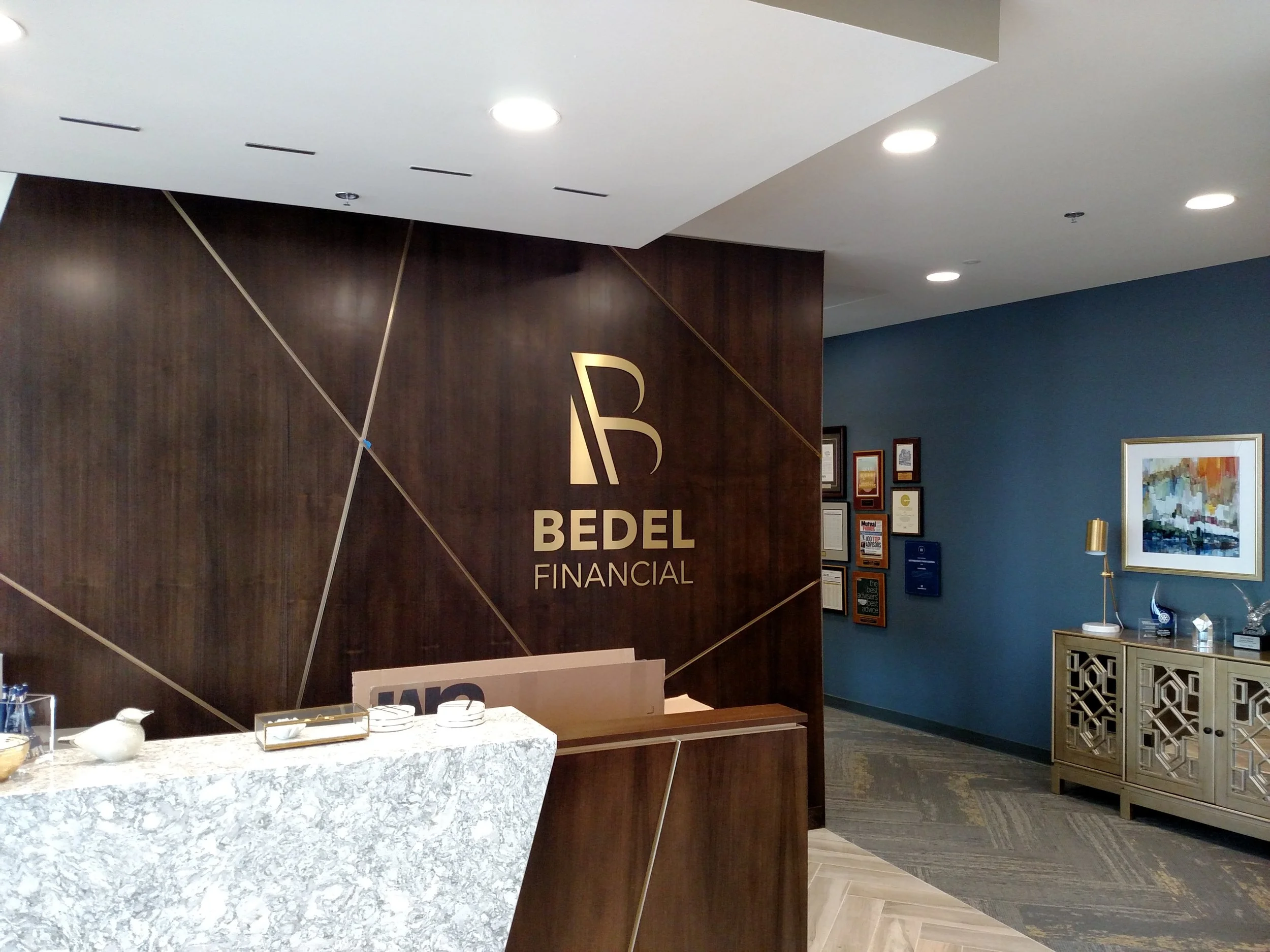 Interior of Bedel Financial office with a reception desk, dark wood wall with logo, blue wall with framed certificates and colorful artwork, and decorative cabinet.