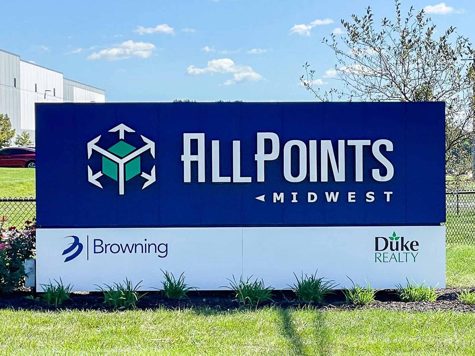Signboard displaying AllPoints Midwest and logos for Browning and Duke Realty with a blue background and a green and white geometric icon, outdoor setting, blue sky with clouds, grassy area with small plants, fence, and trees in background.