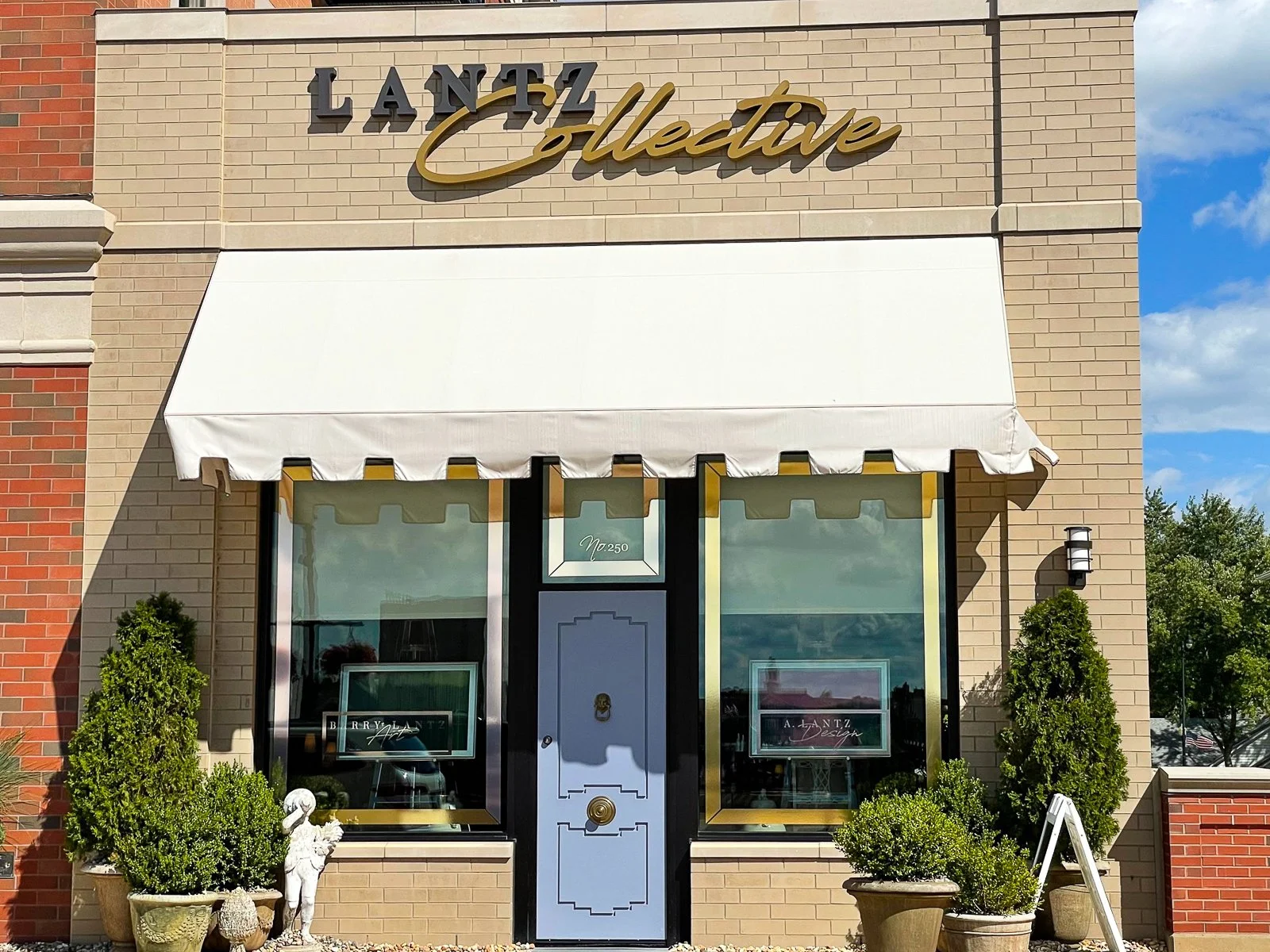 Front view of a storefront with a sign that reads 'LANTZ Collective.' The shop has a white awning, large display windows, and a light blue door in the center. There are potted plants and a decorative statue near the entrance.