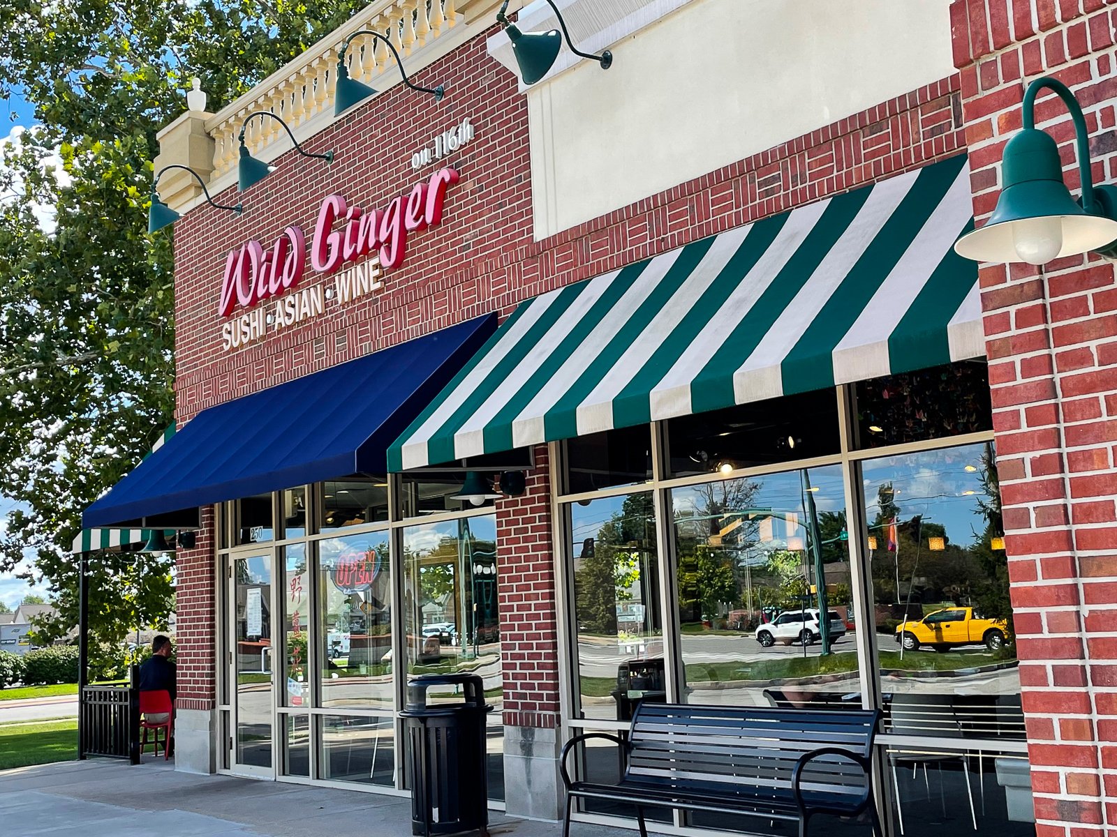 Exterior of Old Ginger restaurant with red brick facade, striped awning, and blue canopy, displaying signs for sushi, Asian cuisine, and wine.