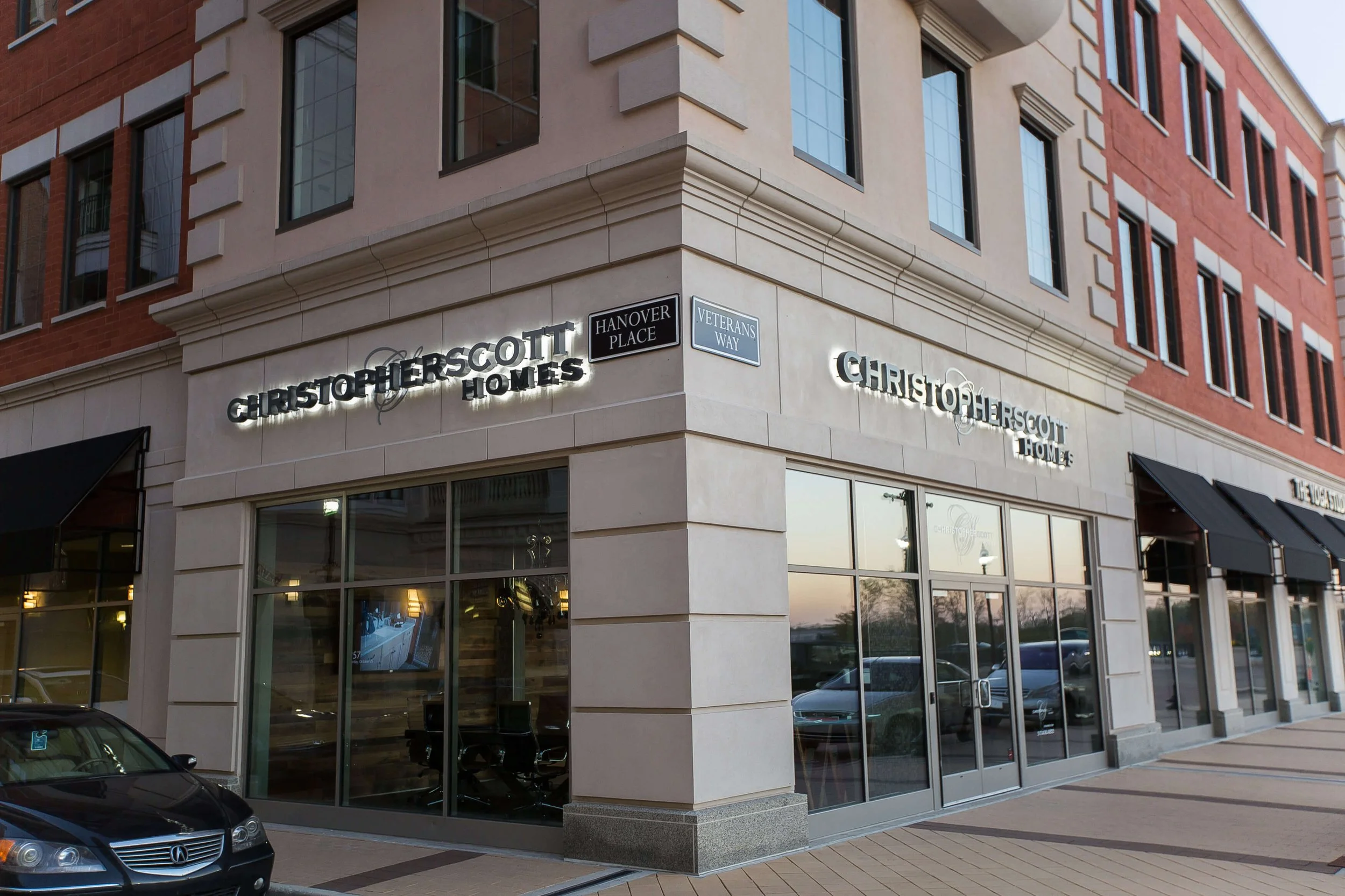 Corner of a multi-story building with signage for Christopher Homes and street signs for Hanover Place and Veterans Way, with large glass windows showing the interior and parked cars outside.