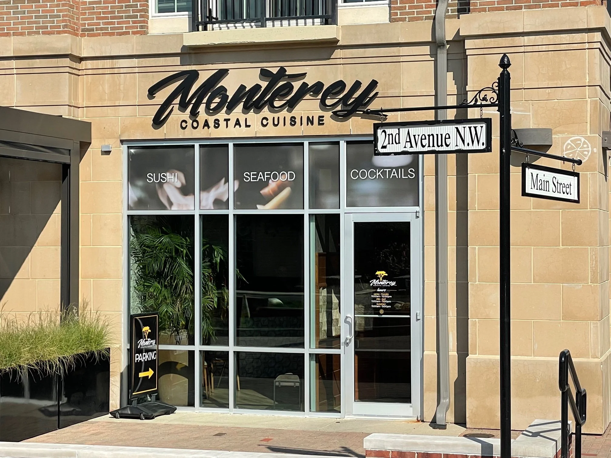 Exterior of Monterey Coastal Cuisine restaurant on 2nd Avenue NW, featuring large windows with signs for sushi, seafood, and cocktails, and street signs for 2nd Avenue NW and Main Street.