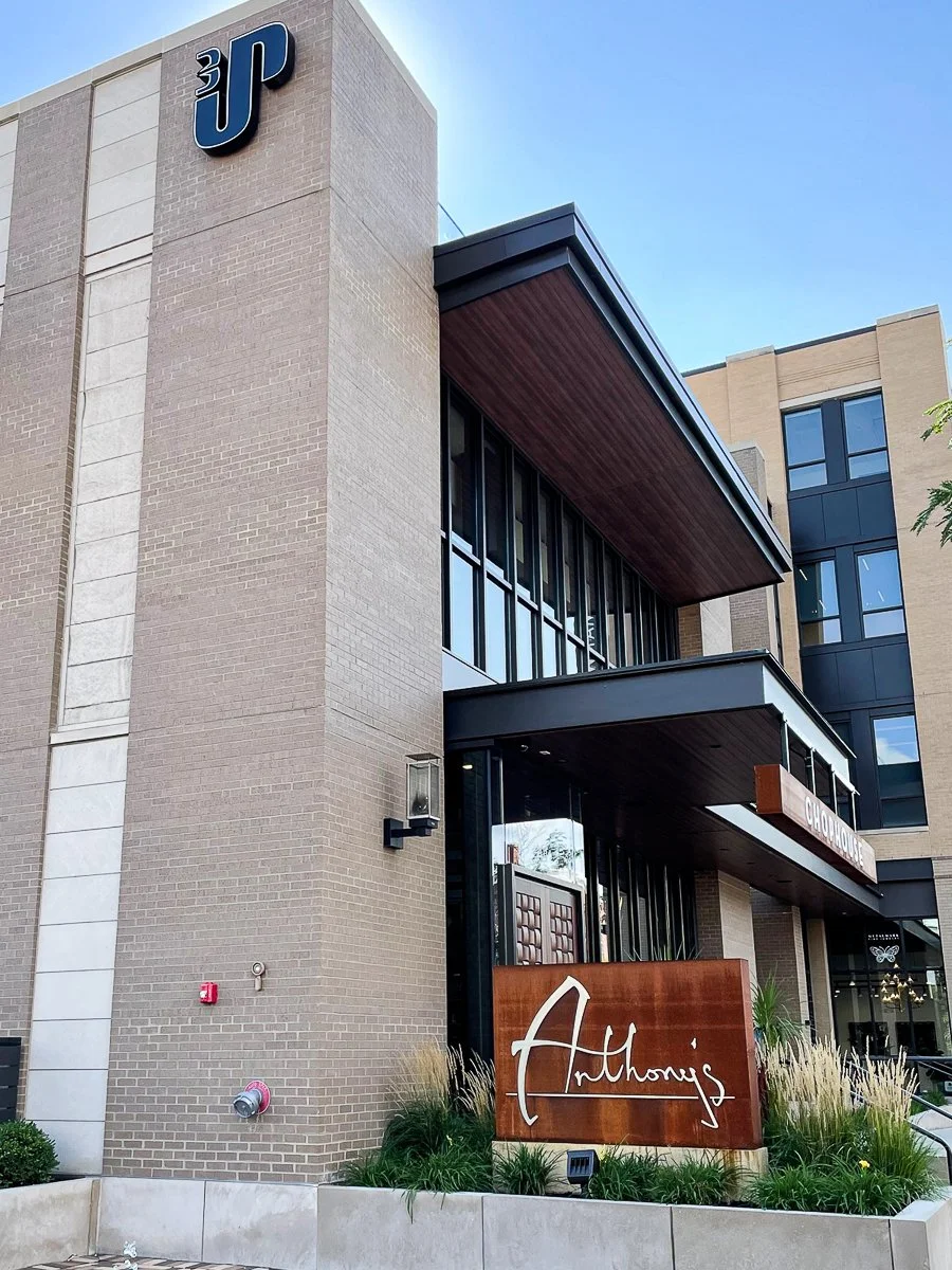 Exterior view of a modern multi-story building with a large sign reading "Anthony's" in front of greenery, and a small sign for a chocolate store above.