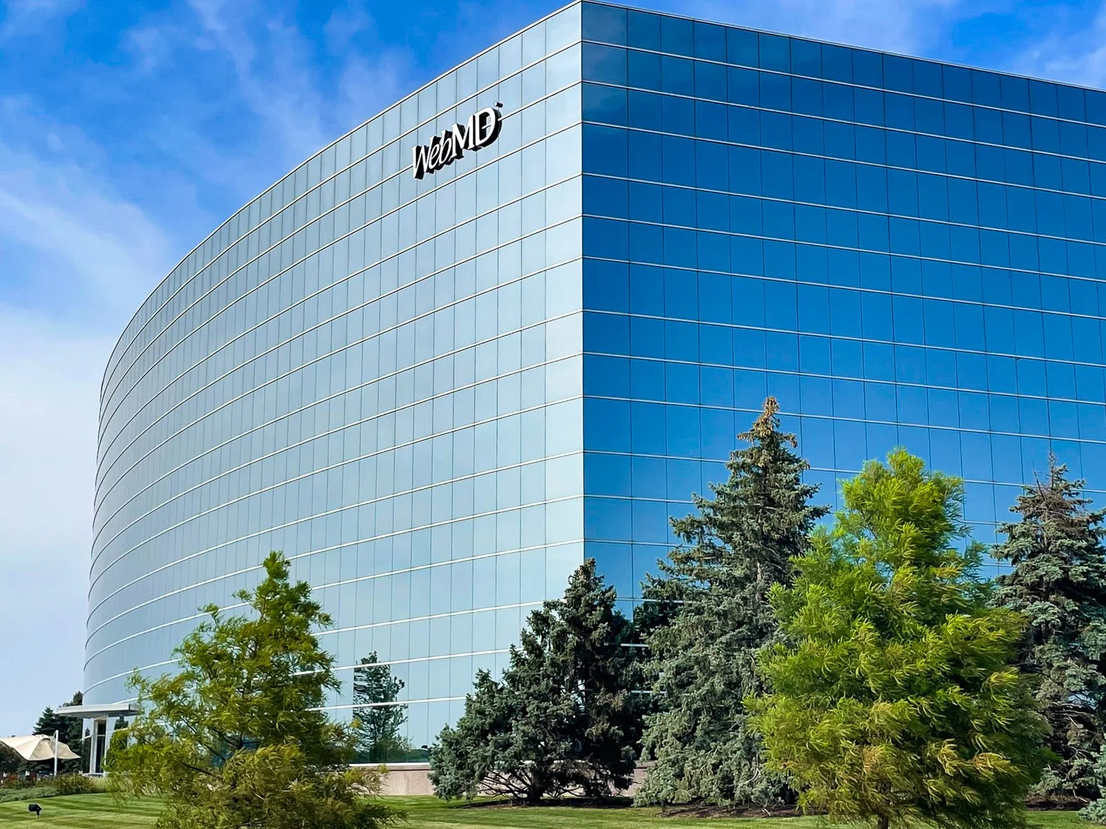 Blue glass building with the WebMD logo at the top, surrounded by green trees and grass, under a partly cloudy sky.