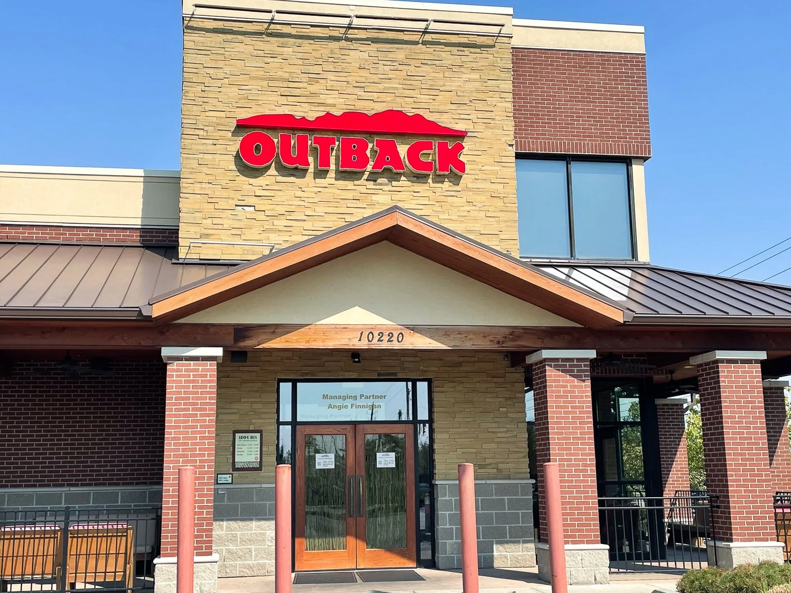 Exterior of an Outback restaurant with a brick and stone facade, red Outback sign, wooden accents, a covered entrance, and benches outside, under a clear blue sky.