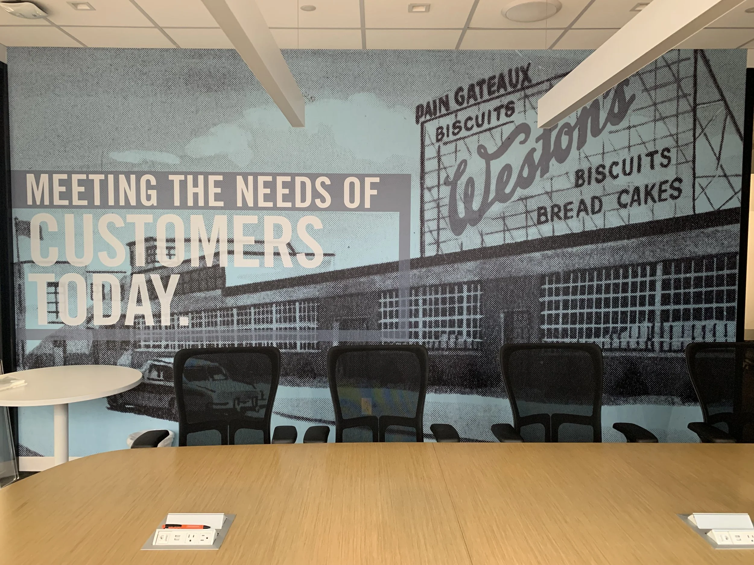 Office conference room with a large mural on the wall featuring a bakery storefront with signs for 'Dain Gateaux,' 'Biscuits,' 'Bread Cakes,' and the word 'Leaston.' A large wooden table with outlets and several chairs in front of the mural.