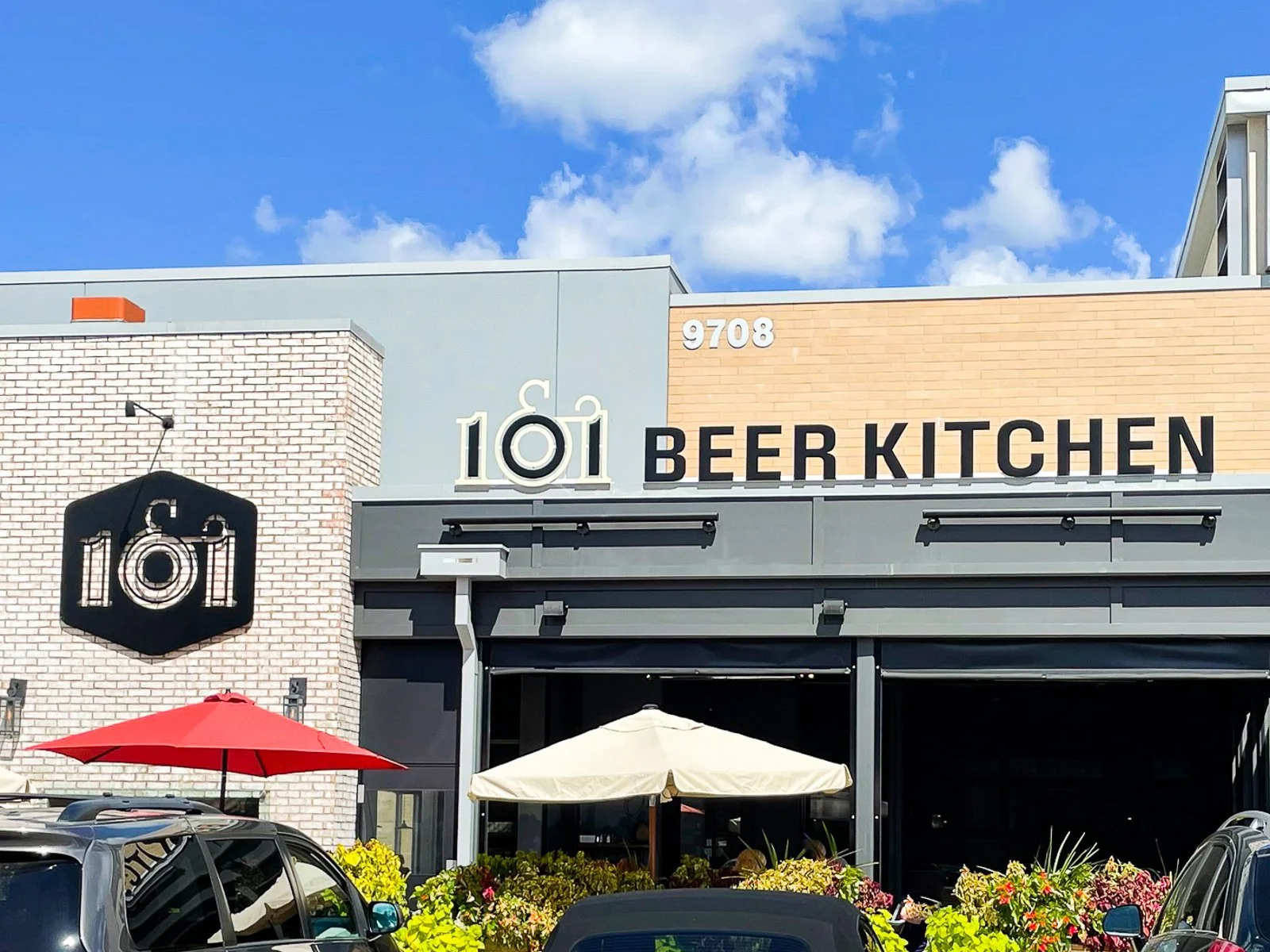 Exterior of a building with a sign reading '101 Beer Kitchen' and the address '9708'. The facade features brick and metal paneling, with an outdoor seating area that has umbrellas and colorful flowers. There are cars parked in front and a large clock