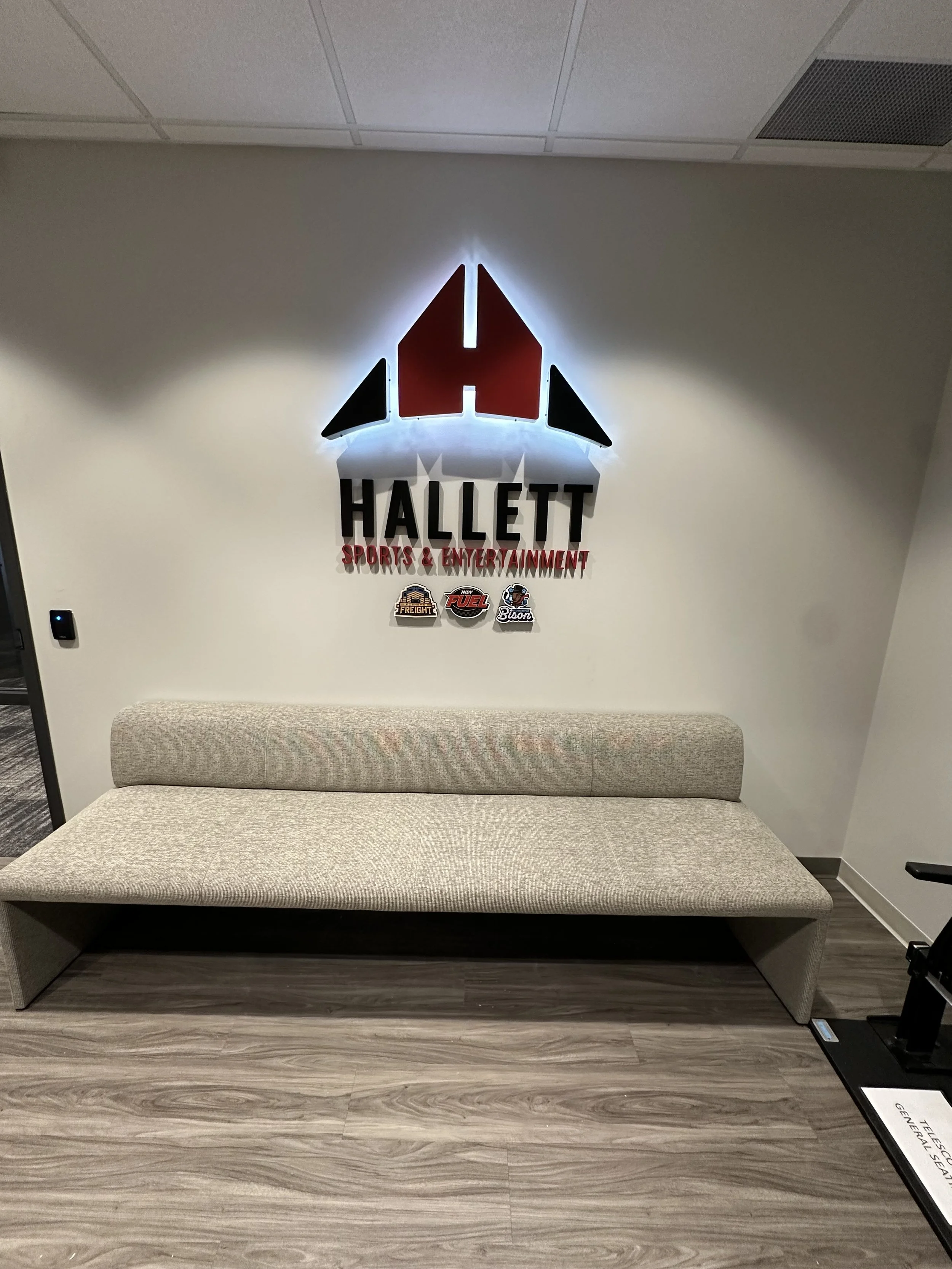 Interior of a waiting area with a beige bench and a wall sign for Hallett Sports & Entertainment, featuring a red and black logo with an arrow shape, and smaller decals of team logos for Freight, Fuel, and Bison.