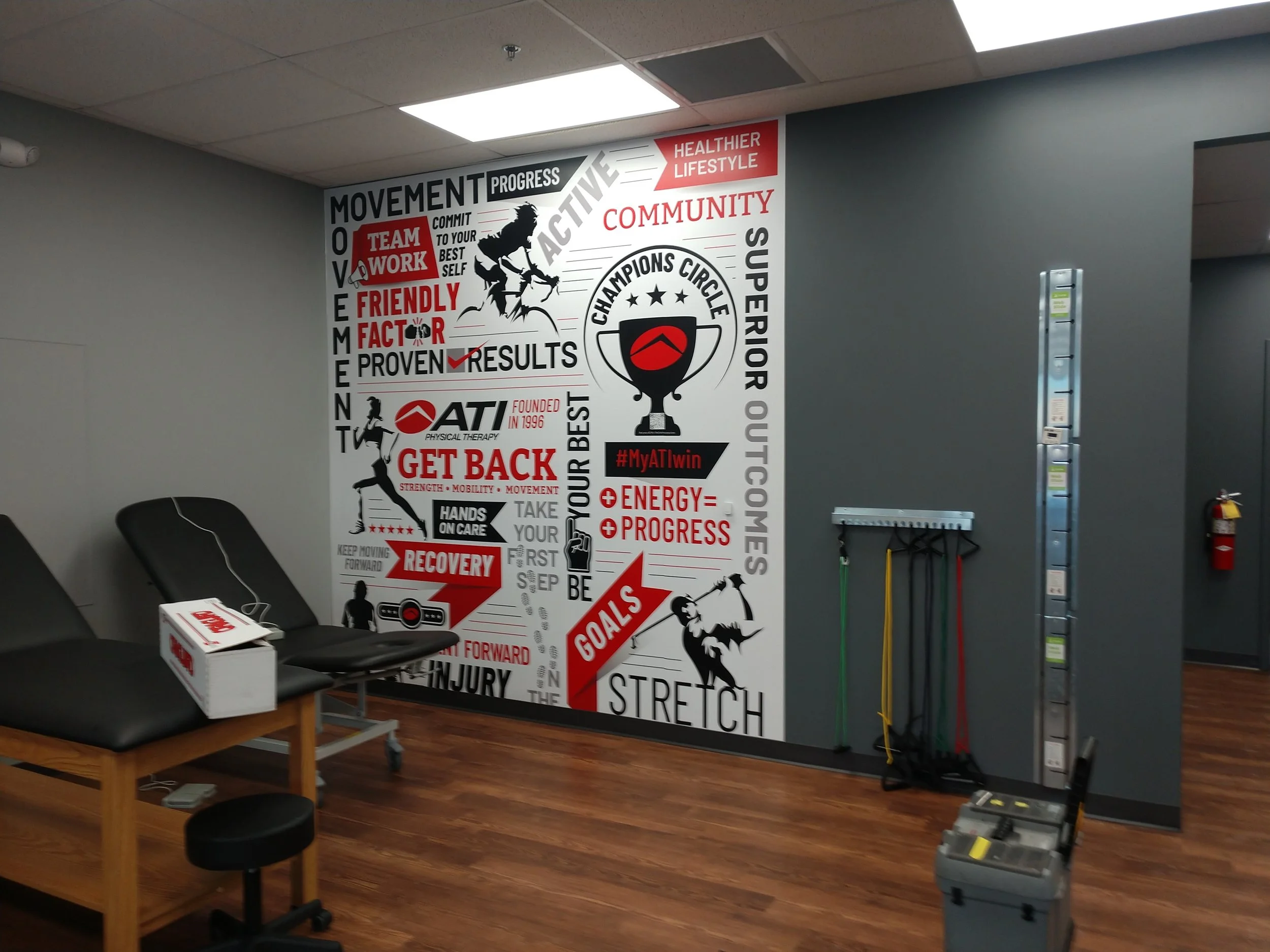 A fitness or physical therapy clinic room with motivational wall art, a black massage or examination table, some exercise resistance bands on a wall rack, and workout equipment on the floor. The wall art features motivational phrases, a trophy, and i