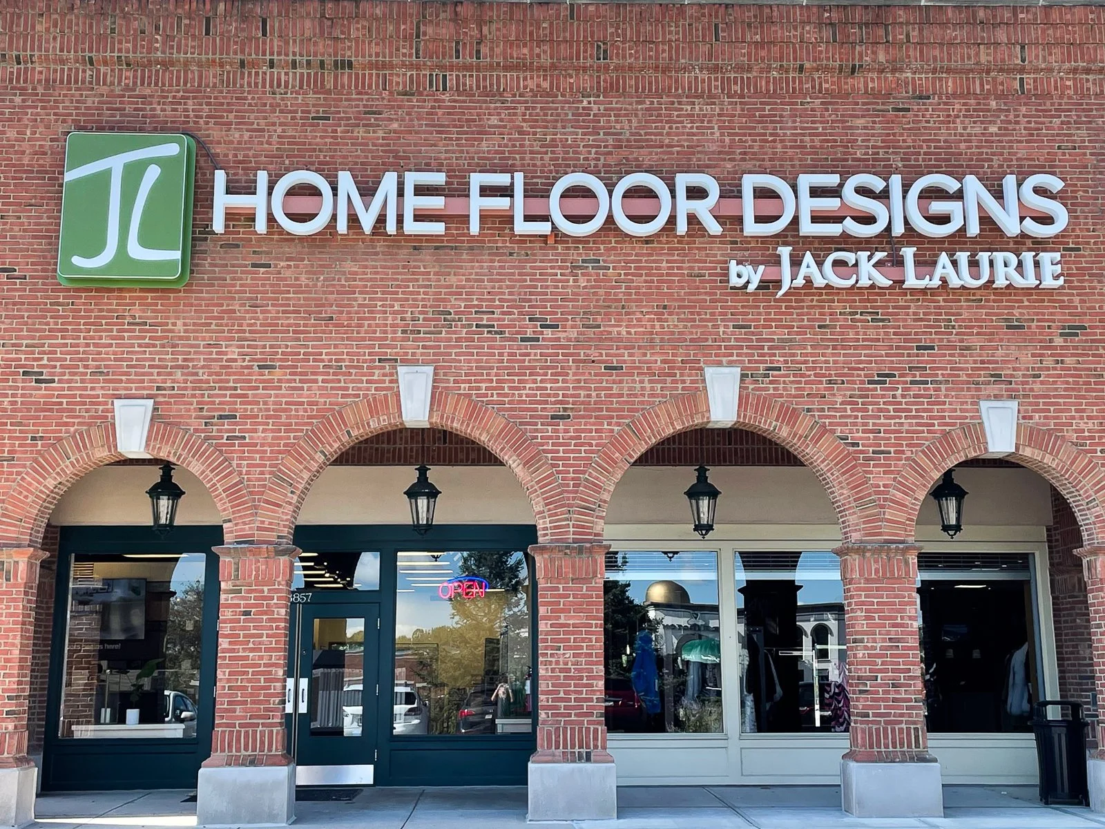 Storefront of Home Floor Designs by Jack Laurie with three brick arches above the entrance and windows. A neon 'Open' sign is visible in the window. The sign on the building includes a green logo with white letters 'JL' and the text 'Home Floor Desig