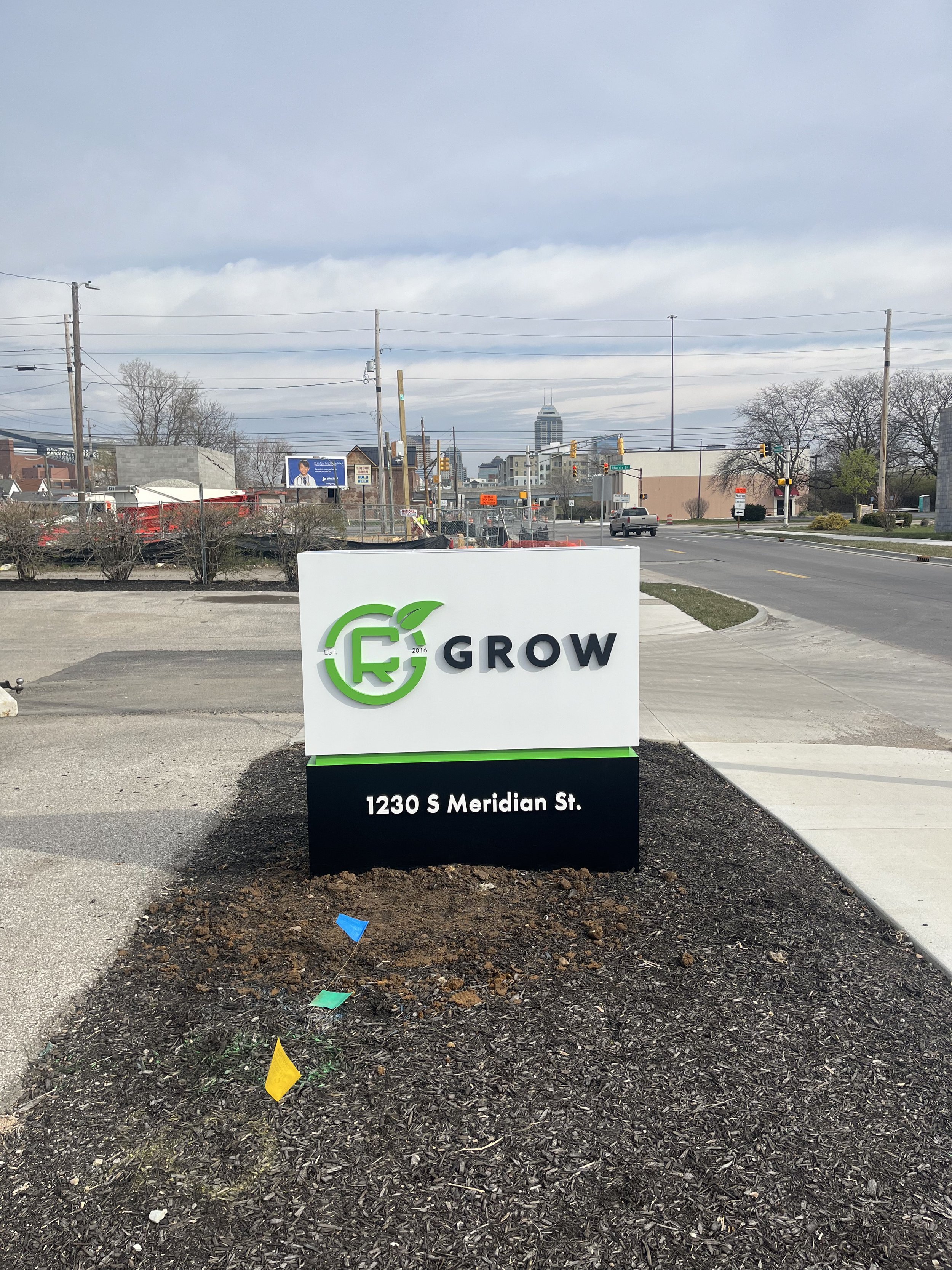 A sign on a sidewalk that reads 'GROW 1230 S Meridian St.' with a logo featuring a green leaf and the letter G, against an urban background with streets, cars, power lines, and a city skyline in the distance.