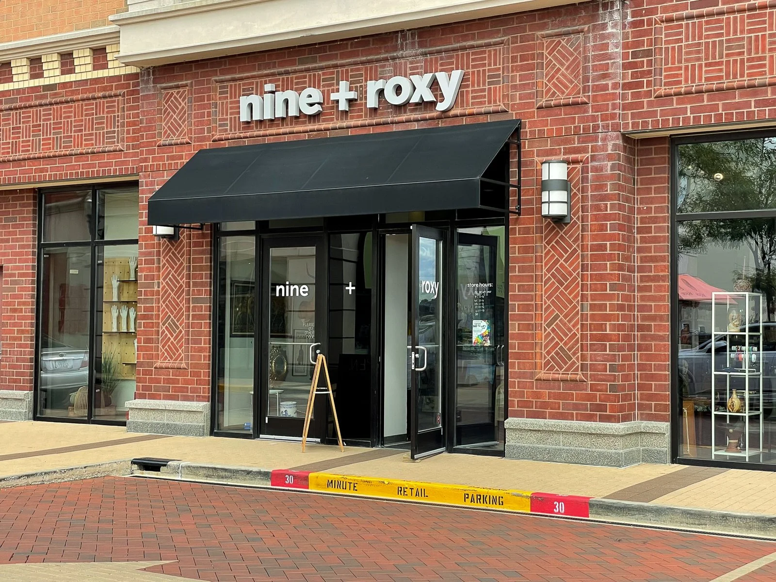 Exterior of a retail store called 'nine + roxy' with a black awning, glass doors, and large windows, part of a red brick building. There is a sidewalk with a yellow and red parking curb in front.