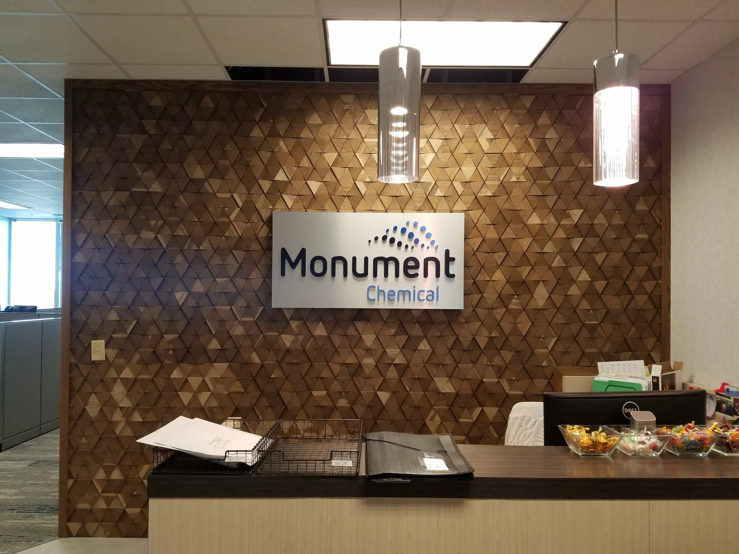 Reception area of Monument Chemical with a wooden textured wall, a sign reading 'Monument Chemical', and a counter with documents, a computer, and bowls of candies.
