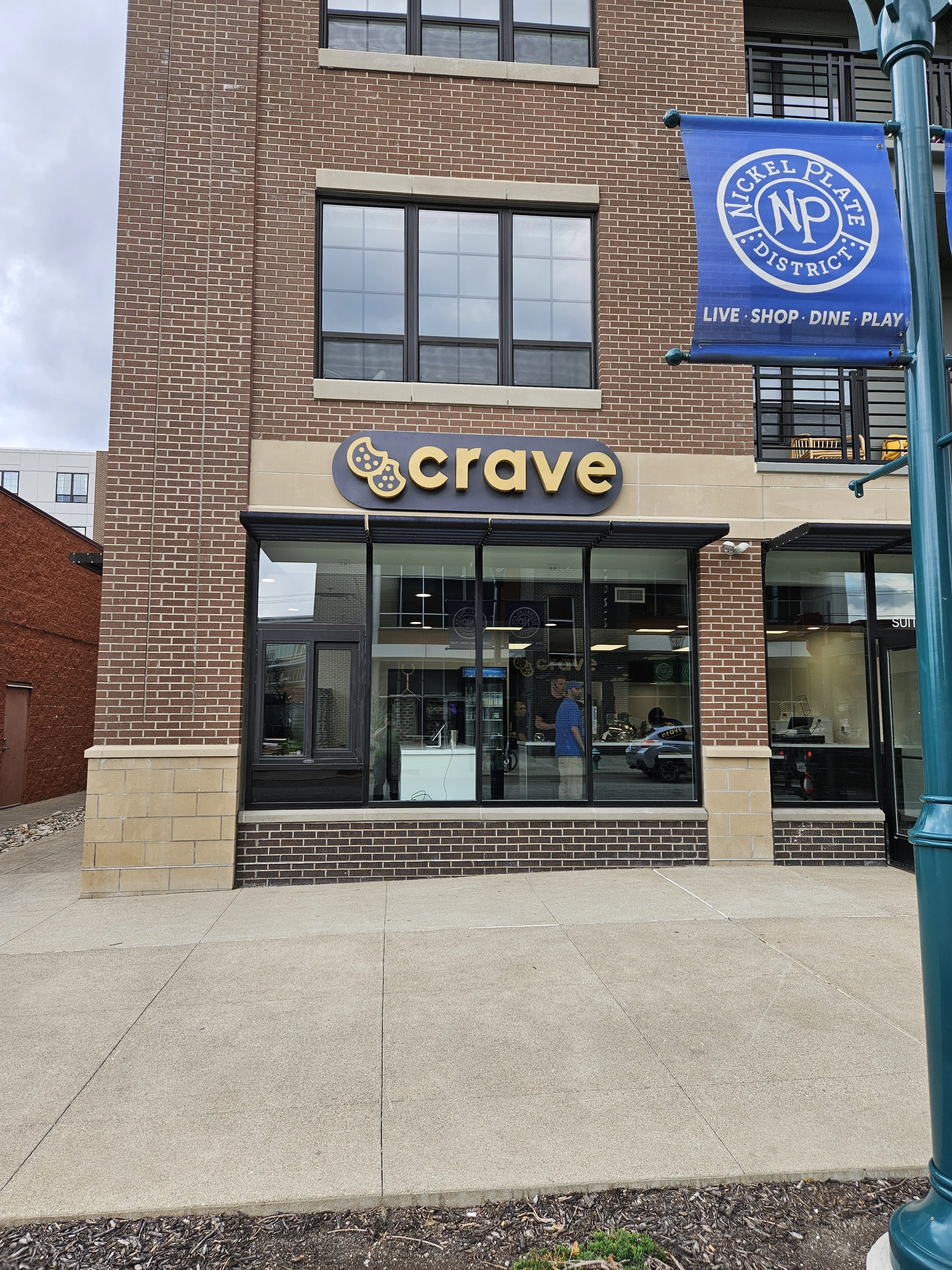  storefront with a sign that says 'crave' and a logo with a cookie, located in a brick building in Nickel Plate District, near a blue banner that reads 'Nickel Plate District Live Shop Dine Play'.