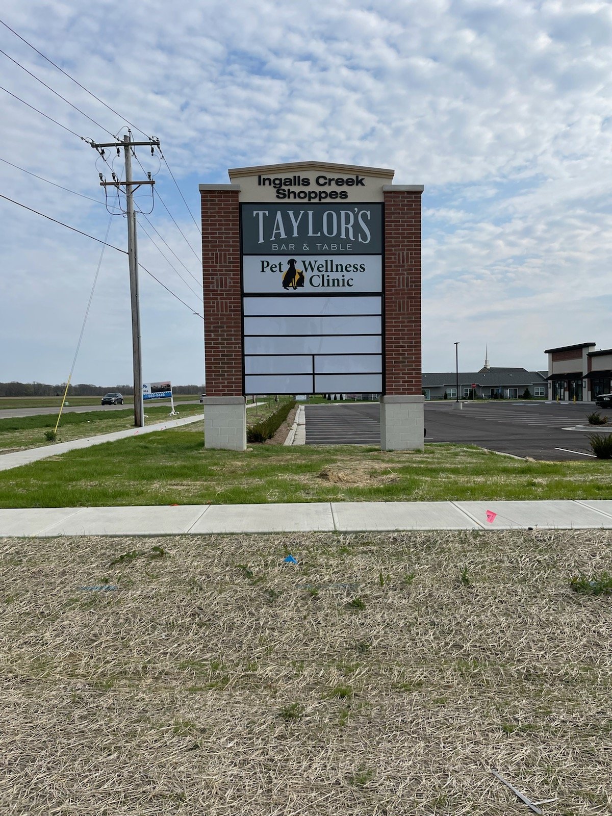 A large sign in front of a shopping plaza with the text 'Ingalls Creek Shoppes' at the top, followed by 'TAYLOR'S BAR & TABLE' and 'Pet Wellness Clinic' with a paw print logo. The parking lot and a few buildings are visible in the background under a 