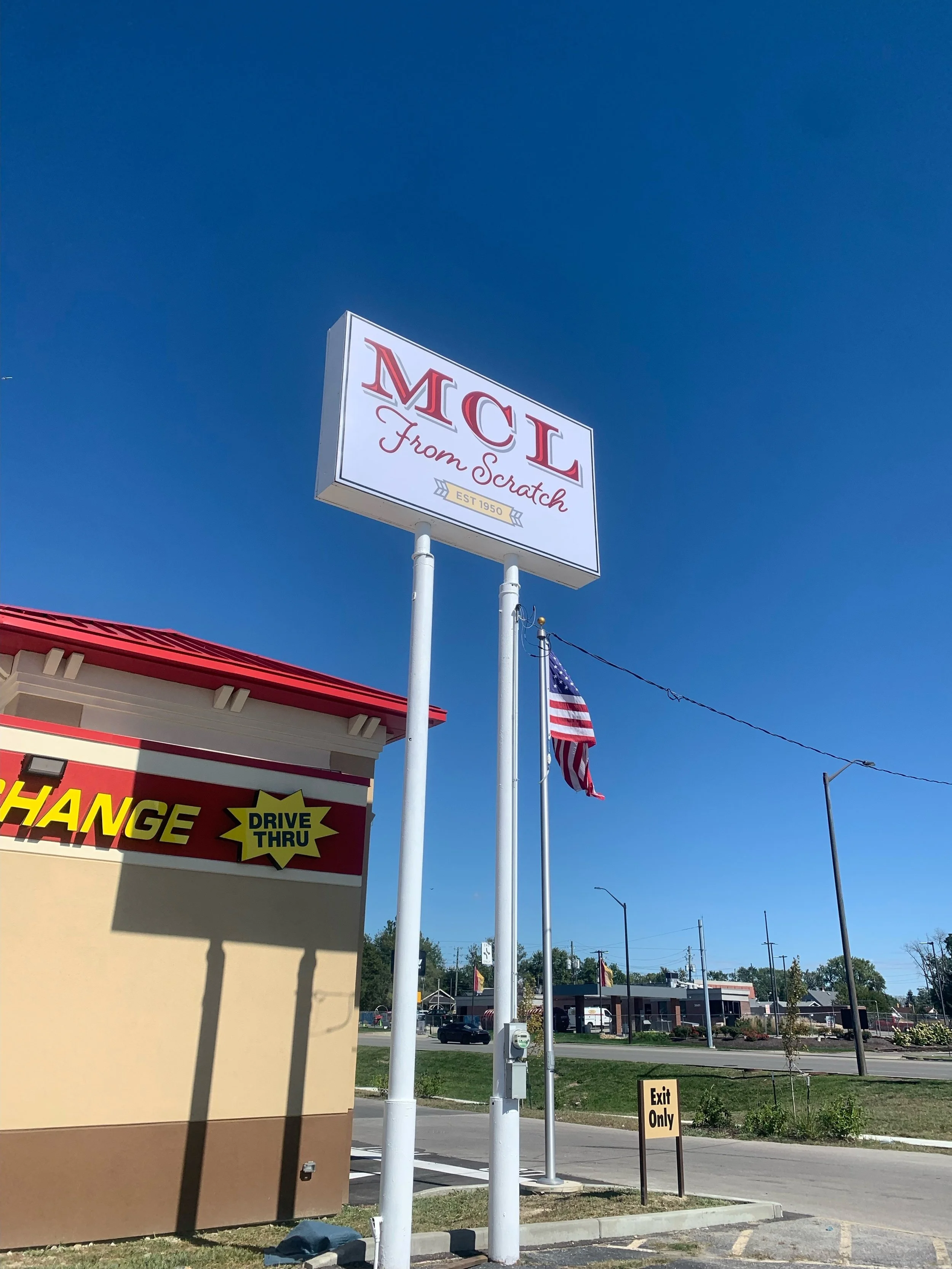 Sign for MCL from Scratch in a parking lot with American flag, drive thru sign, and exit only sign under a clear blue sky.