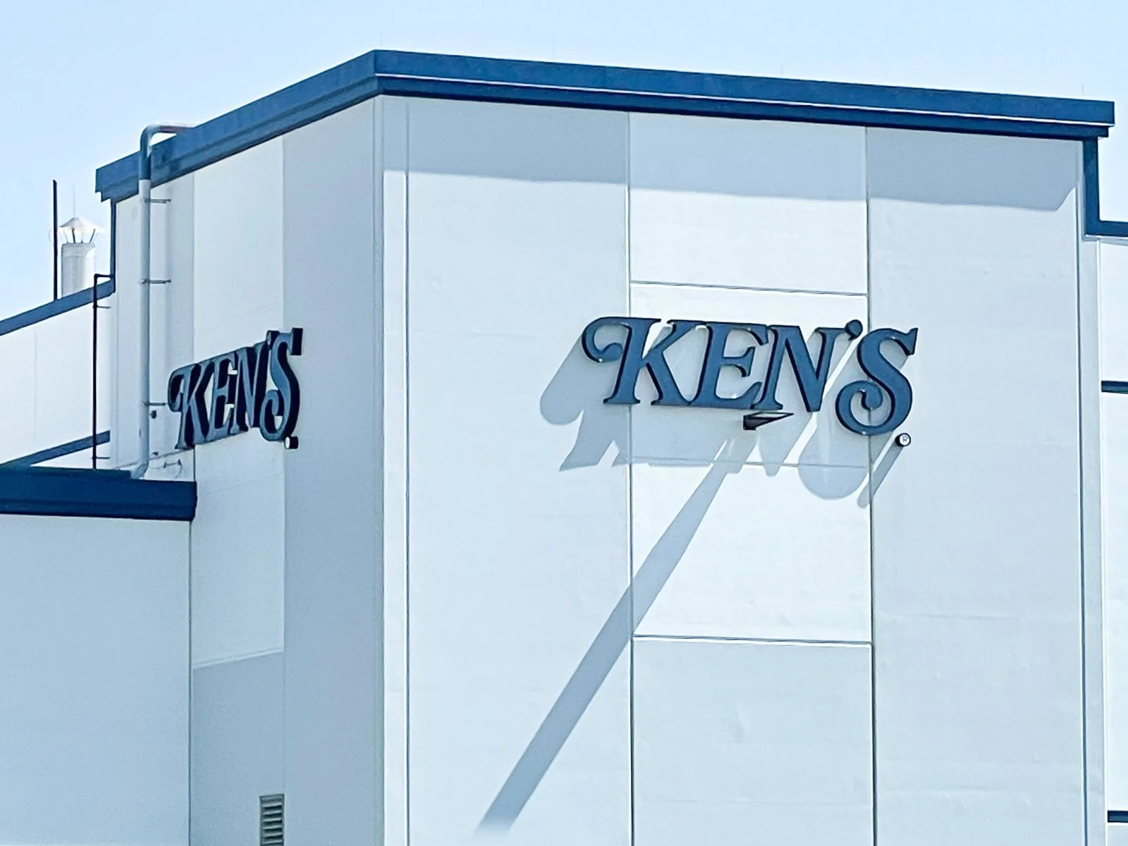 Side of a white building with blue trim, with the word 'KENS' prominently displayed in blue letters on the wall, casting shadows in sunlight.