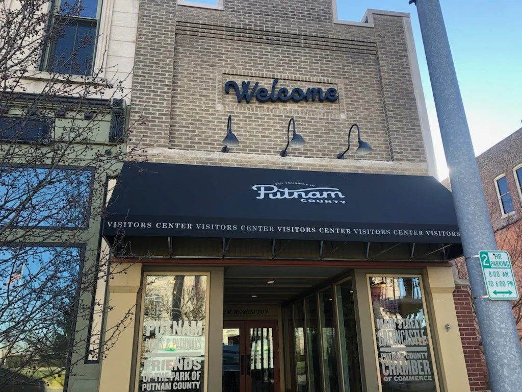 Building with a black awning reading "Putnam County" and windows displaying information, a brick facade, a sign with "Welcome" on the upper wall, and a street sign indicating parking regulations.