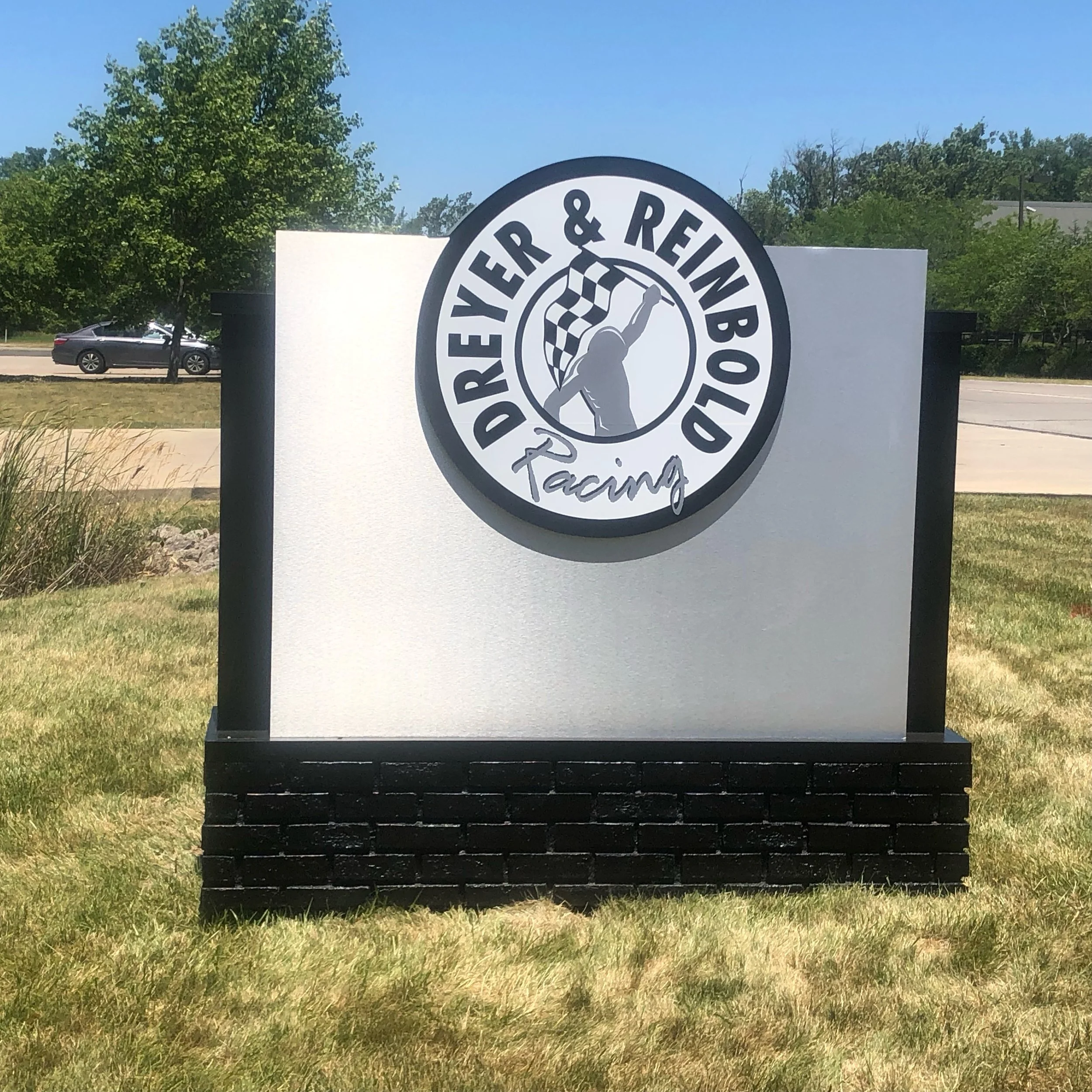 Sign for Dwyer & Reinbold Racing on a grassy area with trees and a road in the background, featuring a logo with a person holding a checkered flag.