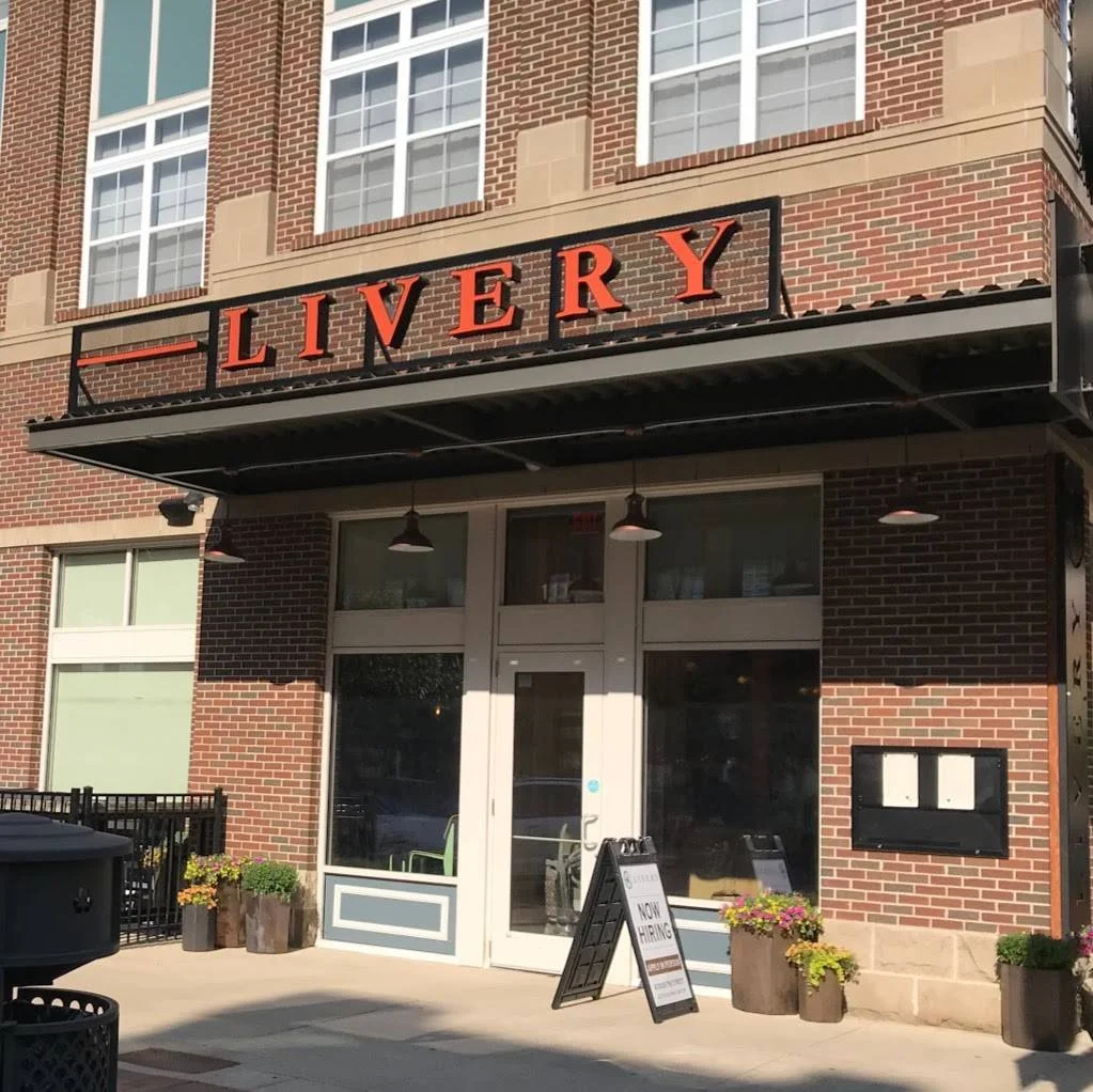 A brick building with a sign that reads 'LIVERY' above the entrance. The entrance has glass doors with some potted plants outside. There is also a chalkboard sign with 'NOW HIRING' written on it.