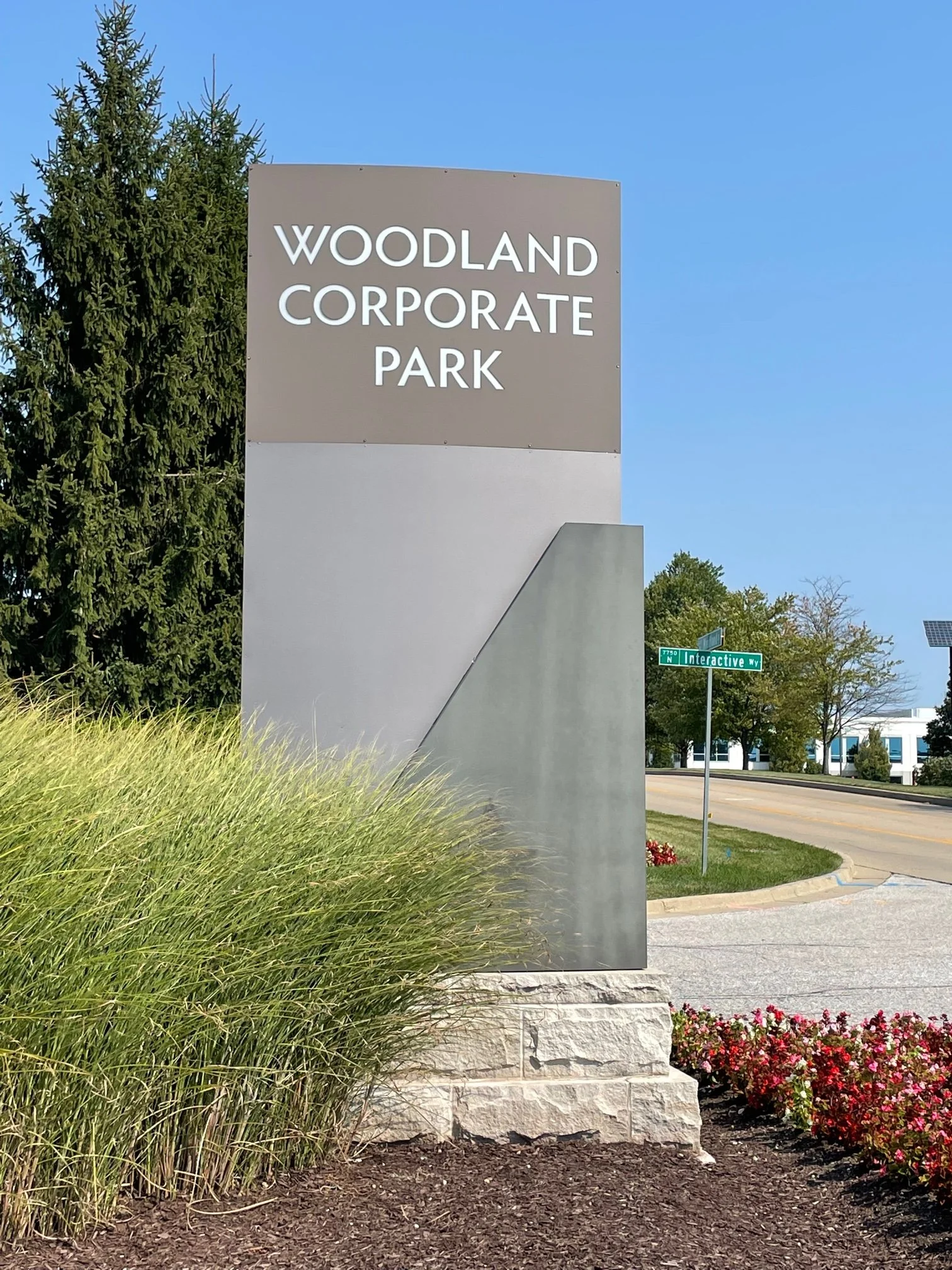 A large sign for Woodland Corporate Park positioned on a landscaped roadside with tall grass and flowers, displaying the name of the business park on a bright day with clear blue sky.