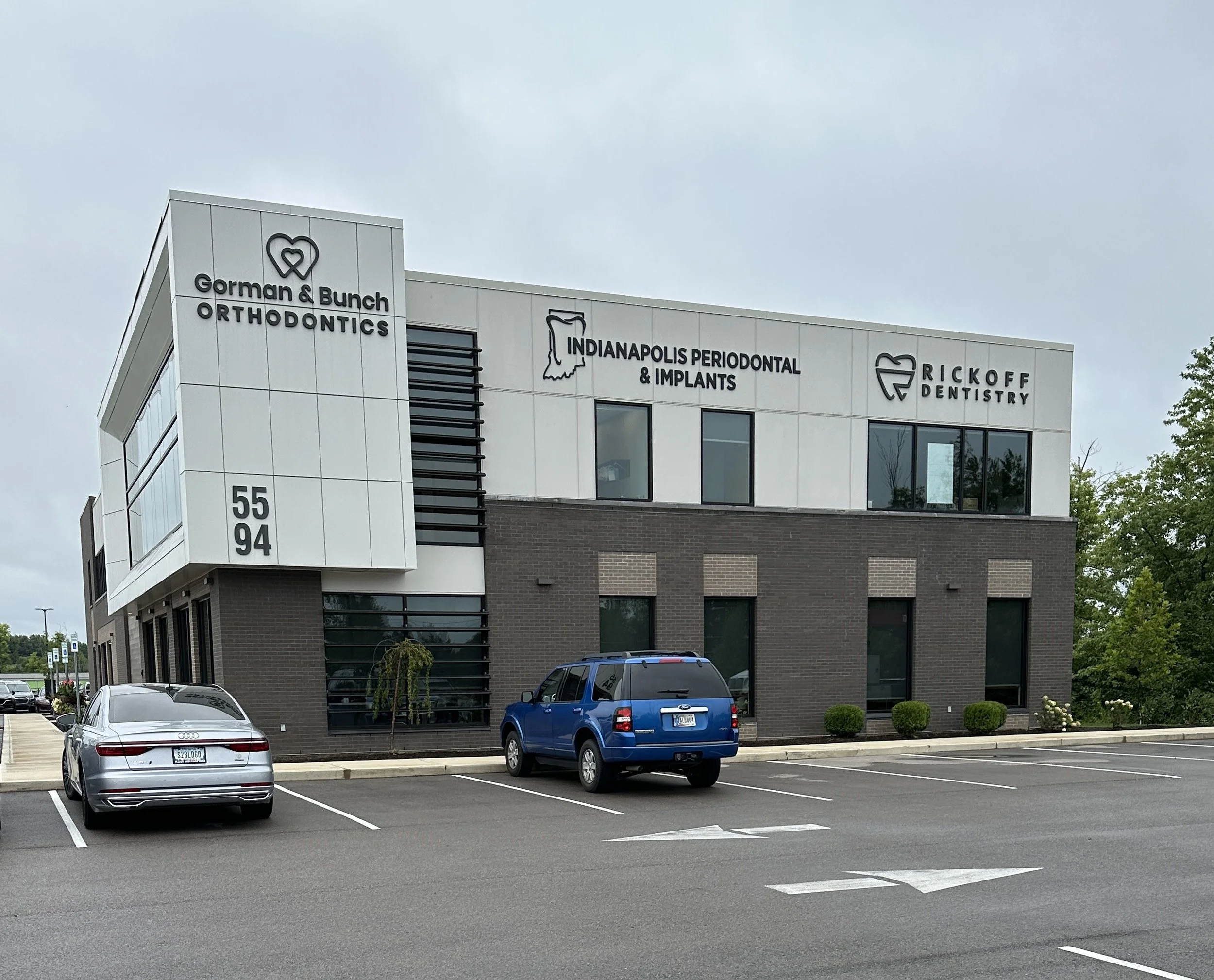 Exterior of a modern multi-tenant dental and orthodontics building with several business signs, including Gorman & Bunch Orthodontics, Indianapolis Periodontal & Implants, and Rickoff Dentistry, with a parking lot in the foreground containing a few p