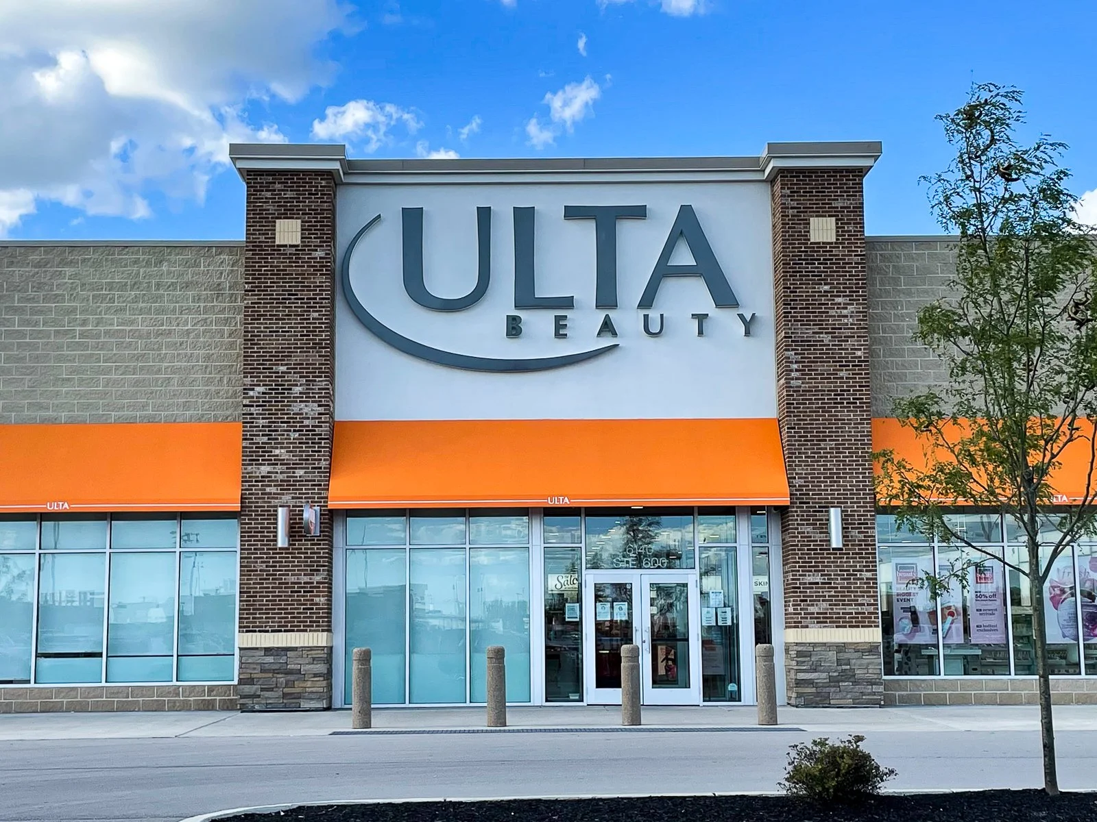 Exterior view of a ULTA Beauty store with large sign on the building, orange awnings, glass doors and windows, and a small tree in front.
