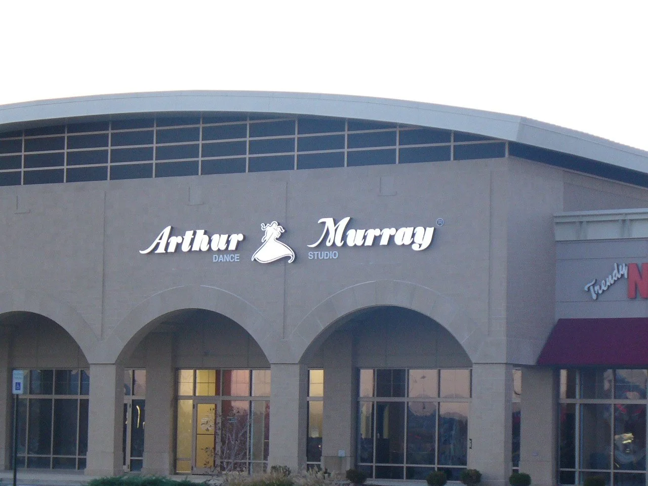 The storefront of Arthur Murray Dance Studio with its sign on the wall, part of a shopping center with arched windows and an adjacent store with a red awning.