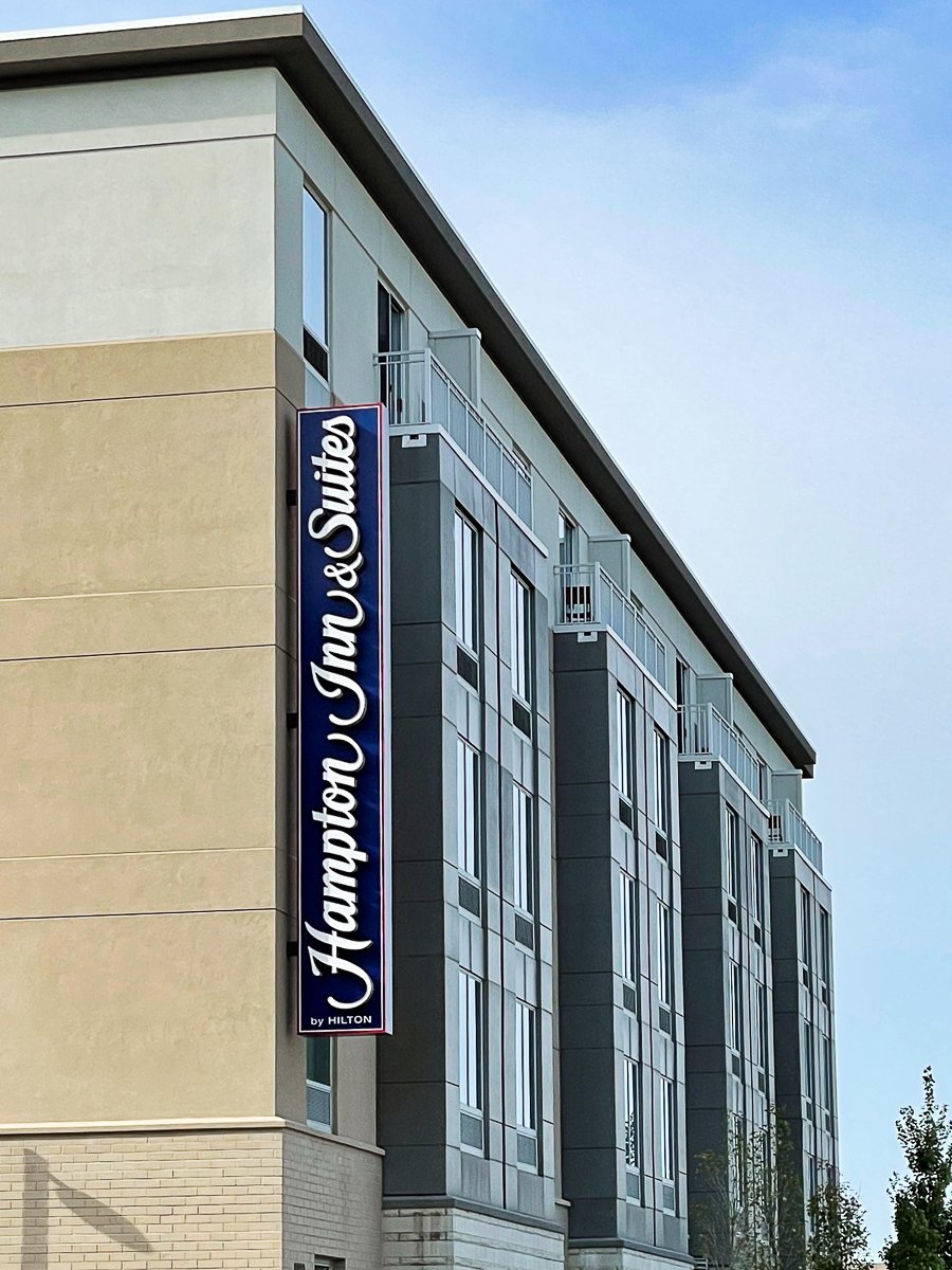 Exterior of a Hilton hotel building with a vertical sign that reads "Hampton Inn & Suites by Hilton" on a beige and gray facade.