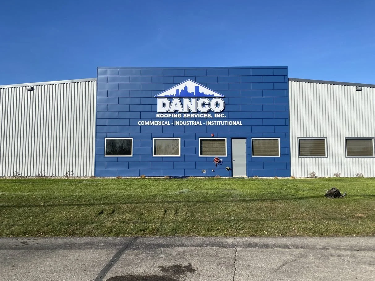 Exterior of a commercial building with a sign for Danco Roofing Services, Inc., featuring a blue facade with the company's logo and windows, under a clear blue sky.