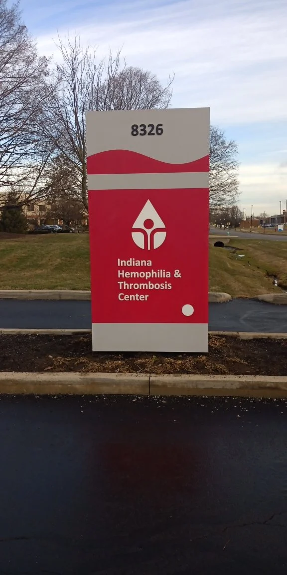 Sign for Indiana Hemophilia & Thrombosis Center with address 8326, featuring a white and red design with a droplet logo, outdoors on a parking lot background.
