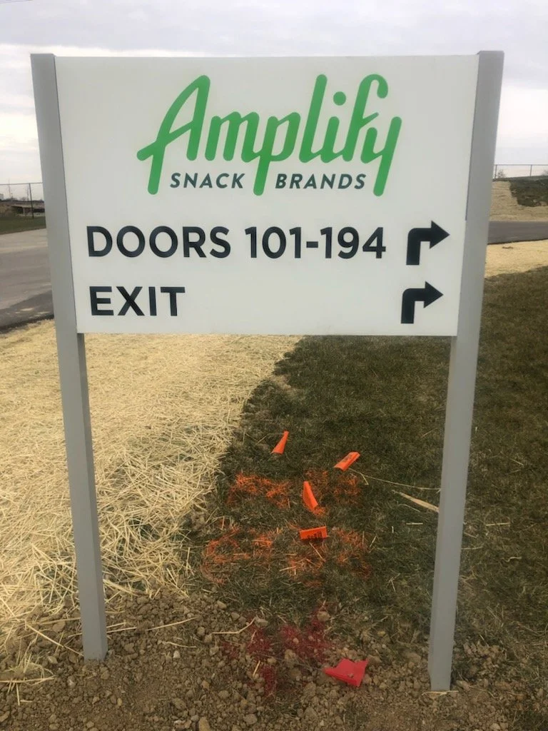 Sign for Amplify Snack Brands directing to doors 101-194 and exit with arrows, and a small orange construction sign and cones on the ground nearby.