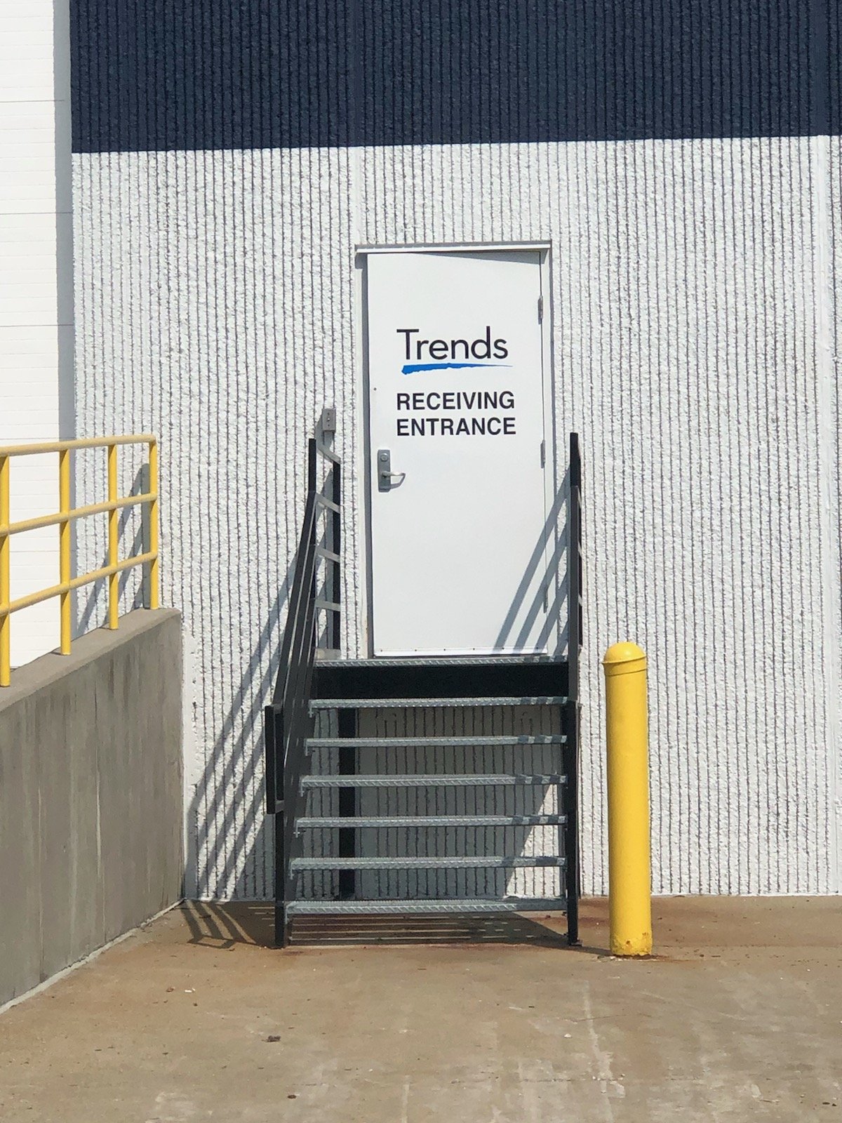A white door labeled 'Trends RECEIVING ENTRANCE' on a building wall, with a black metal staircase leading up to it, a yellow safety bollard on the right, and a yellow railing on the left.