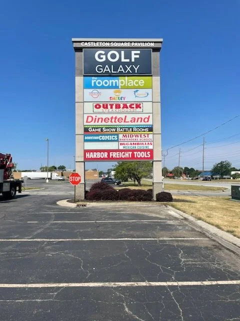 A large outdoor signage board displaying multiple business names, including GOLF GALAXY, room+place, Smiley, OUTBACK, DinetteLand, GAME SHOW BATTLE ROOMS, MID WEST, HARBOR FREIGHT TOOLS, and others, located in a parking lot with a stop sign nearby.