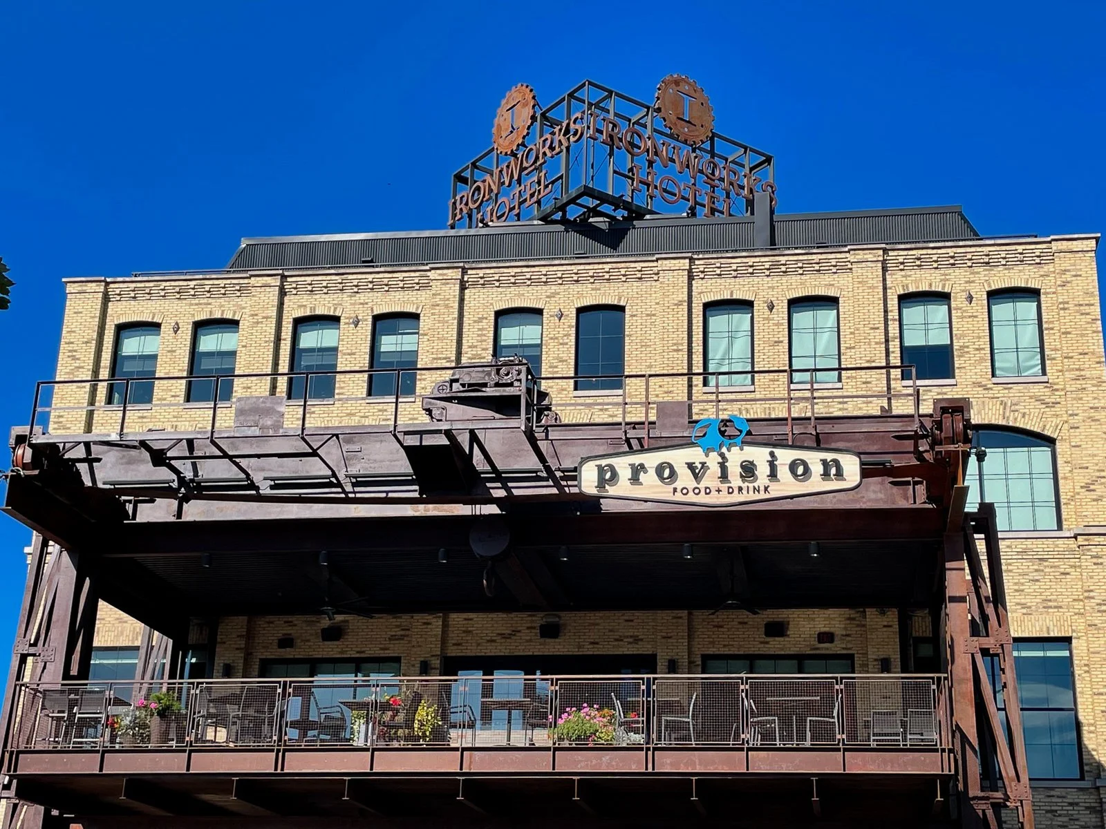 The facade of the Iron Workshop Hotel with a large rooftop sign, and a lower level dining area labeled Providence Food + Drink, featuring outdoor seating with potted plants and chairs.