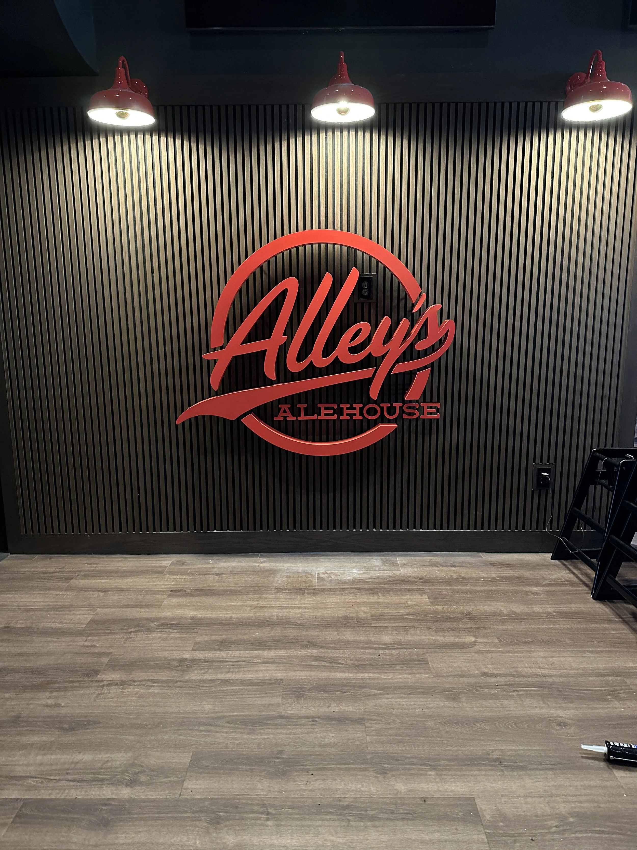 A wall with black vertical slats, three red industrial-style lamps above, and a large red circular sign that reads 'Alley's Alehouse'.
