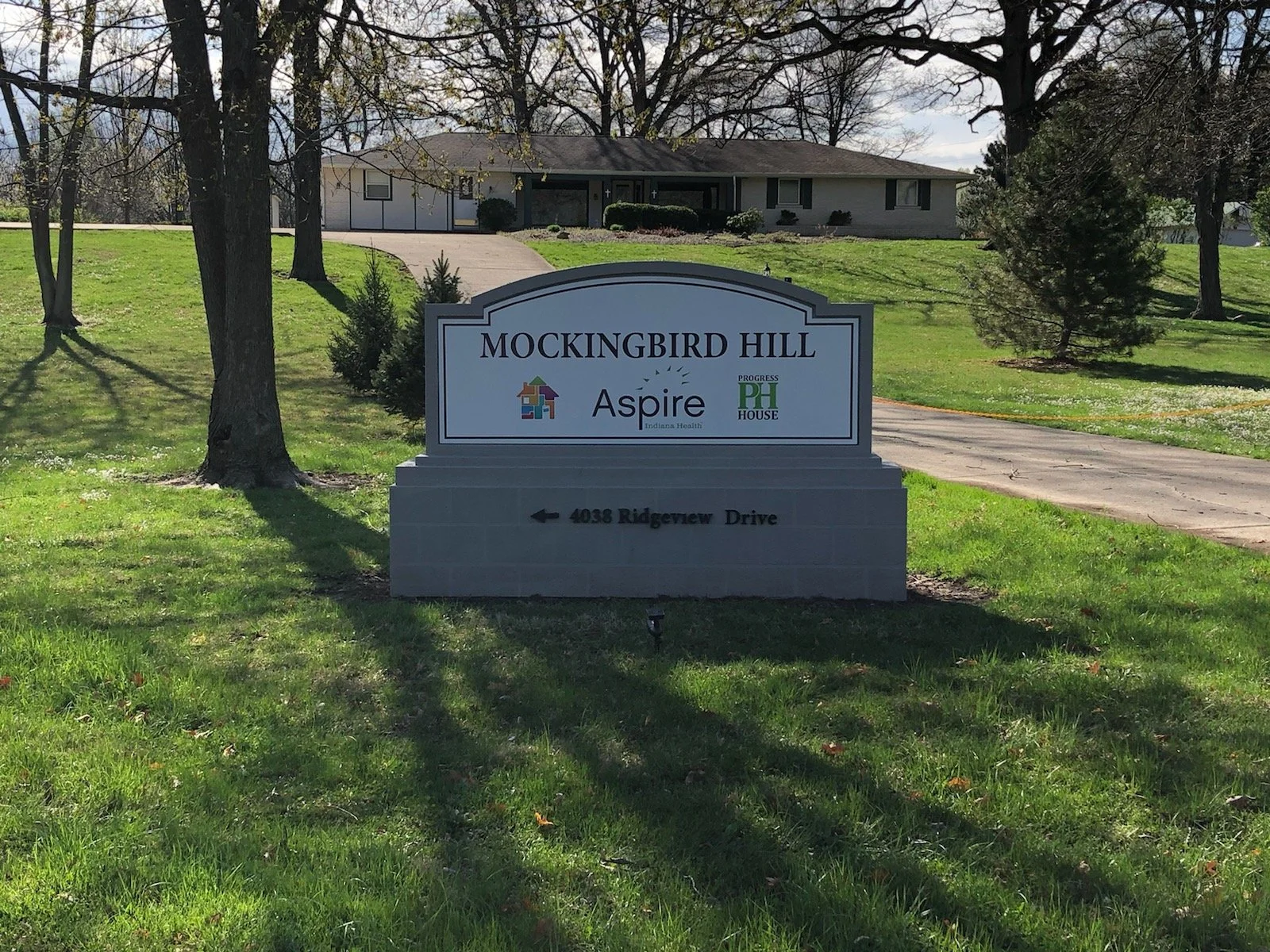Sign for Mockingbird Hill with logos for Aspire Indiana Health and Progress House, and an arrow pointing left with the address 4038 Ridgeview Drive.