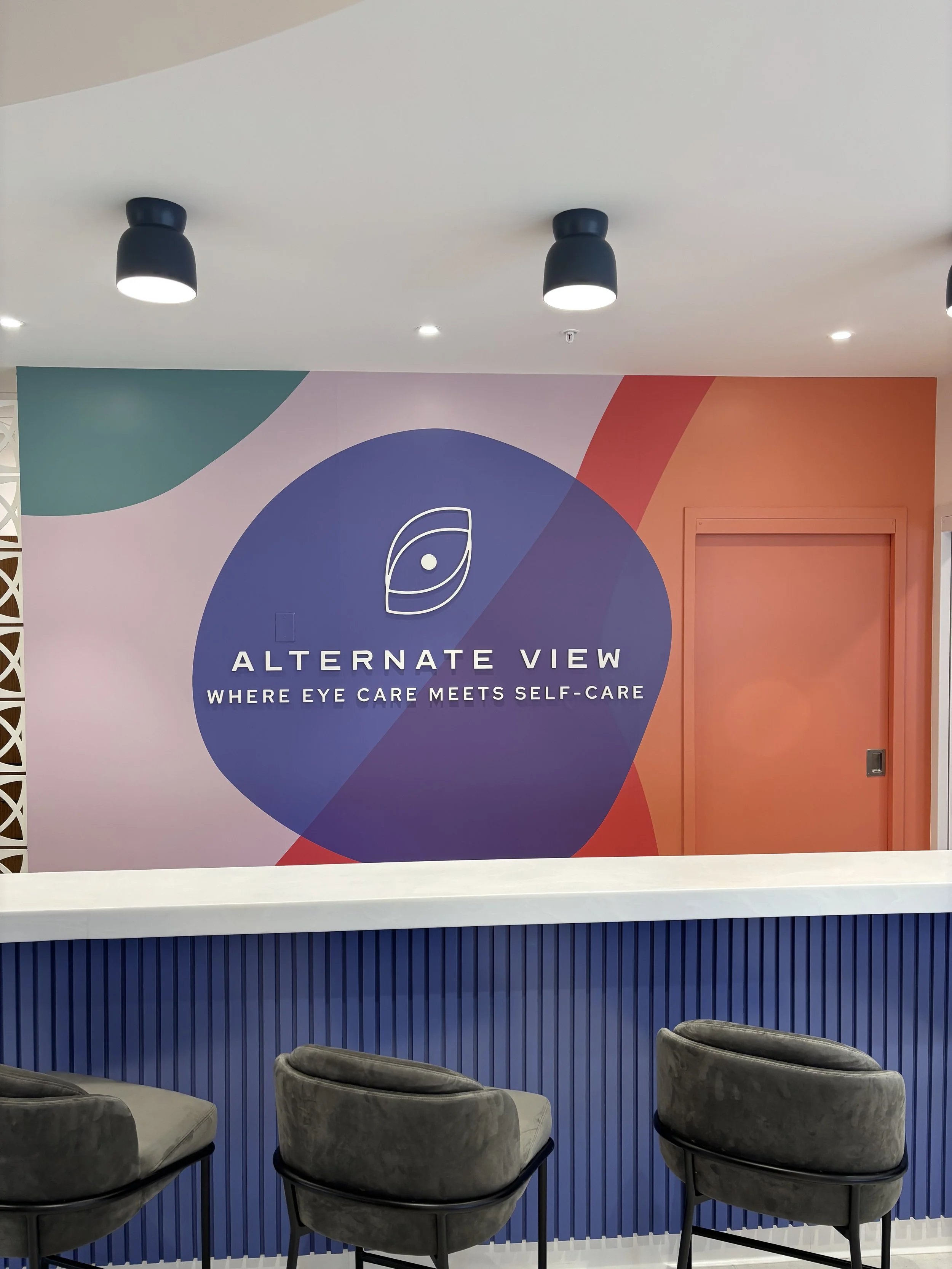 Interior of a modern eye care clinic with a large colorful logo wall that says "Alternate View: Where Eye Care Meets Self-Care." There are three gray chairs in front of the counter.