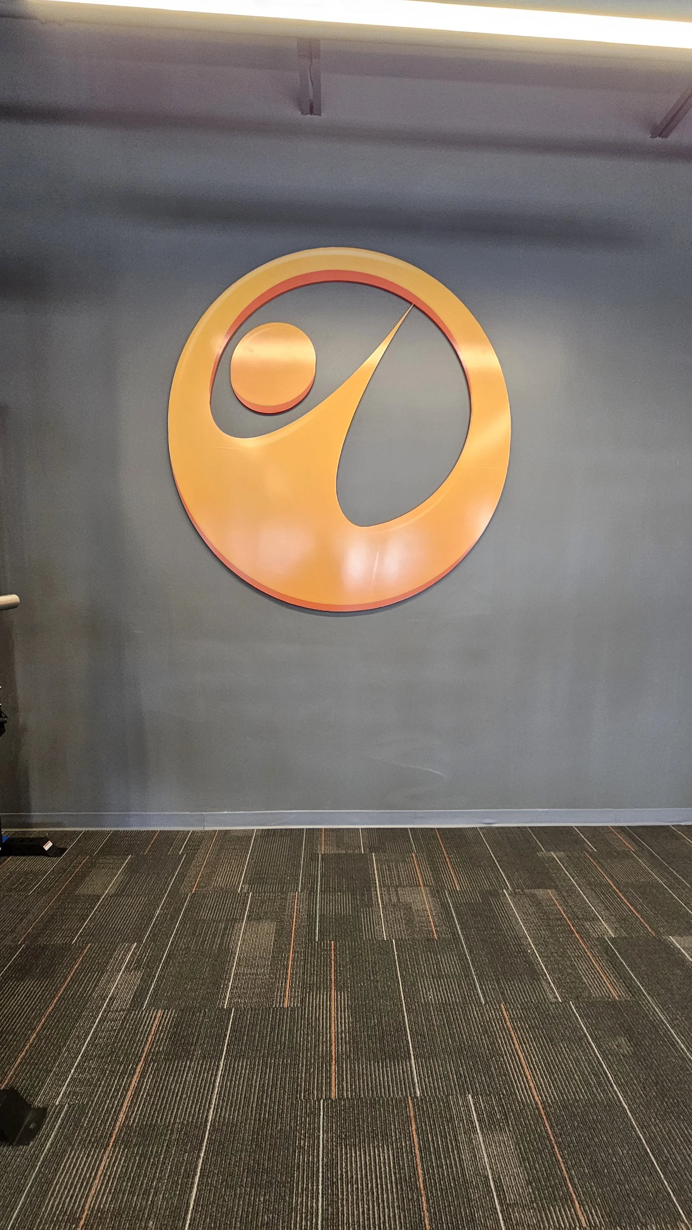 A large orange and red logo on a gray wall featuring a stylized person with an outstretched arm and a dot representing the head.