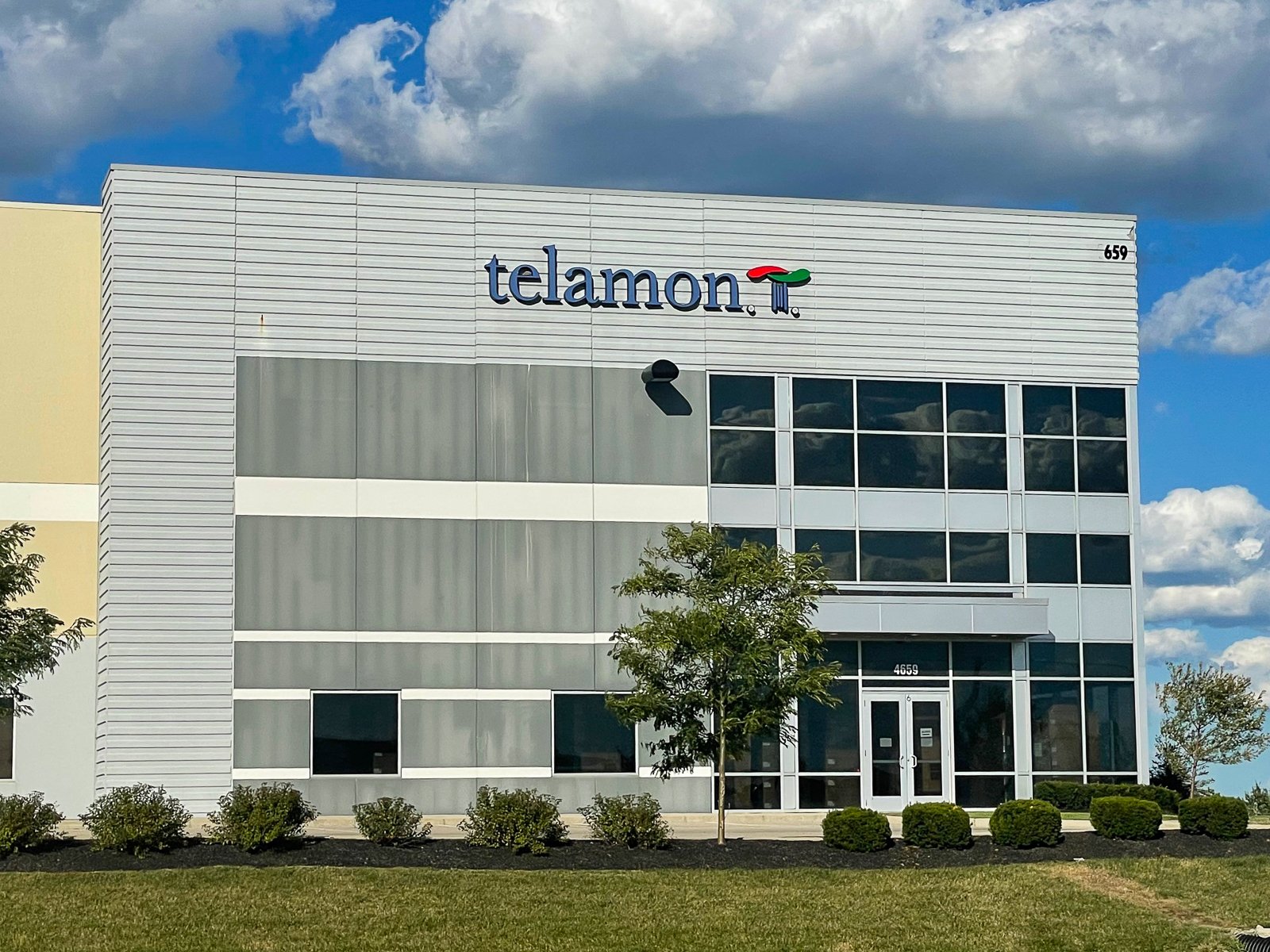 Front view of a modern, three-story office building with glass windows and metal siding, featuring the Telamon logo and an address number 4659, with trees and shrubs in the landscaped front yard.